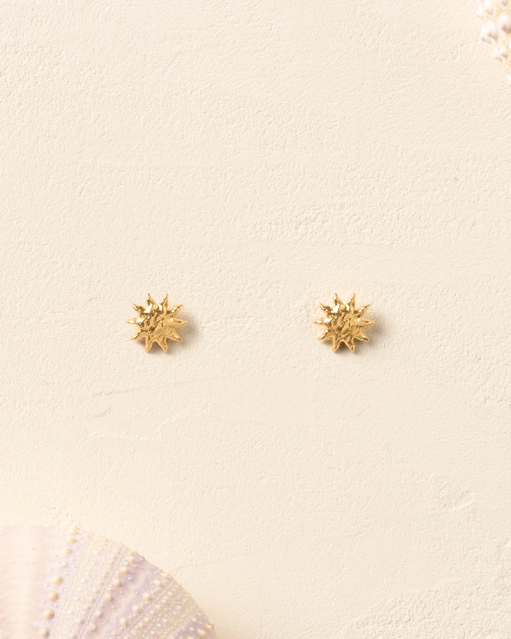 Gold Solé Stud Earrings with sculpted, domed, and hammered sun design on a sandy background.