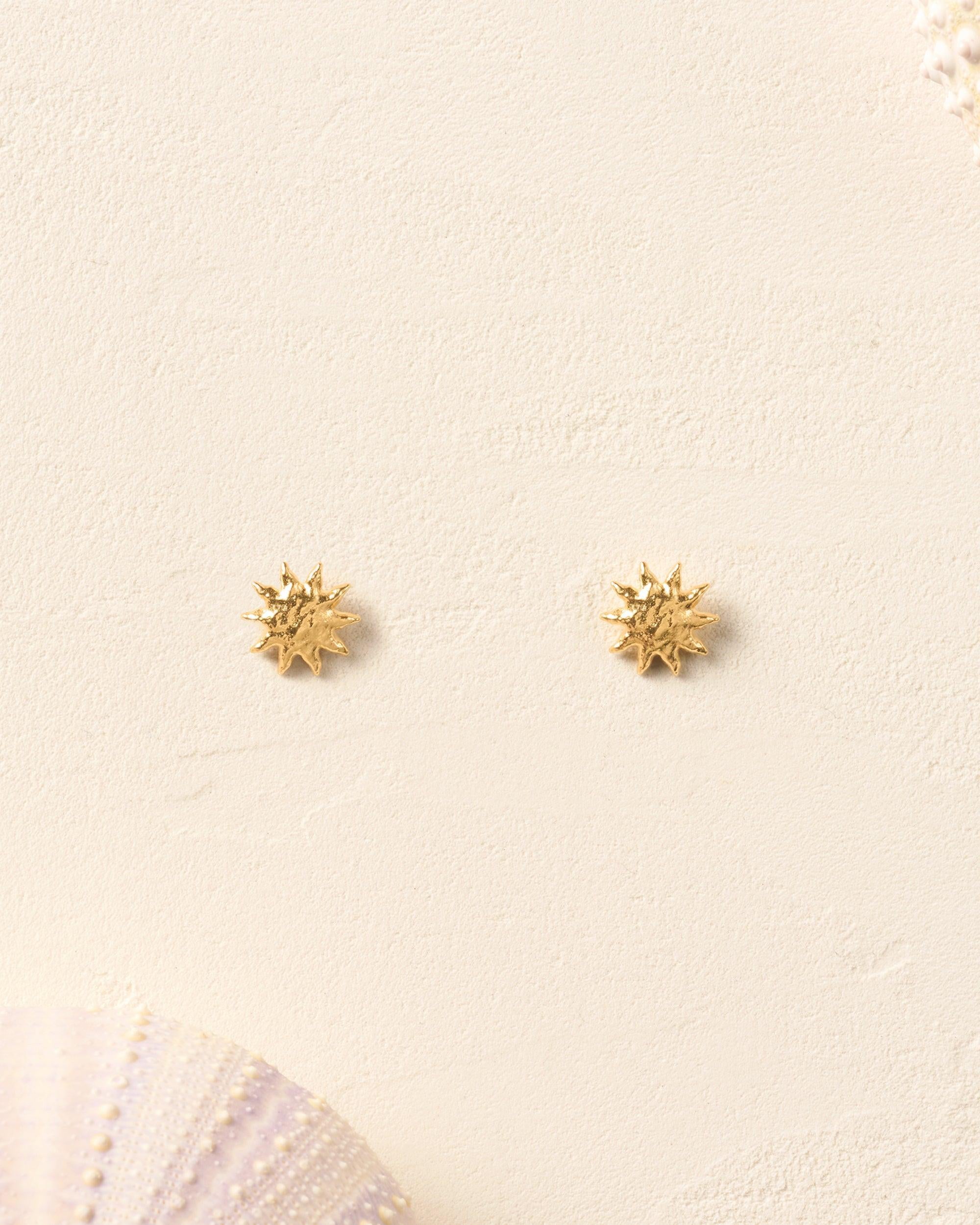 Gold Solé Stud Earrings with sculpted, domed, and hammered sun design on a sandy background.