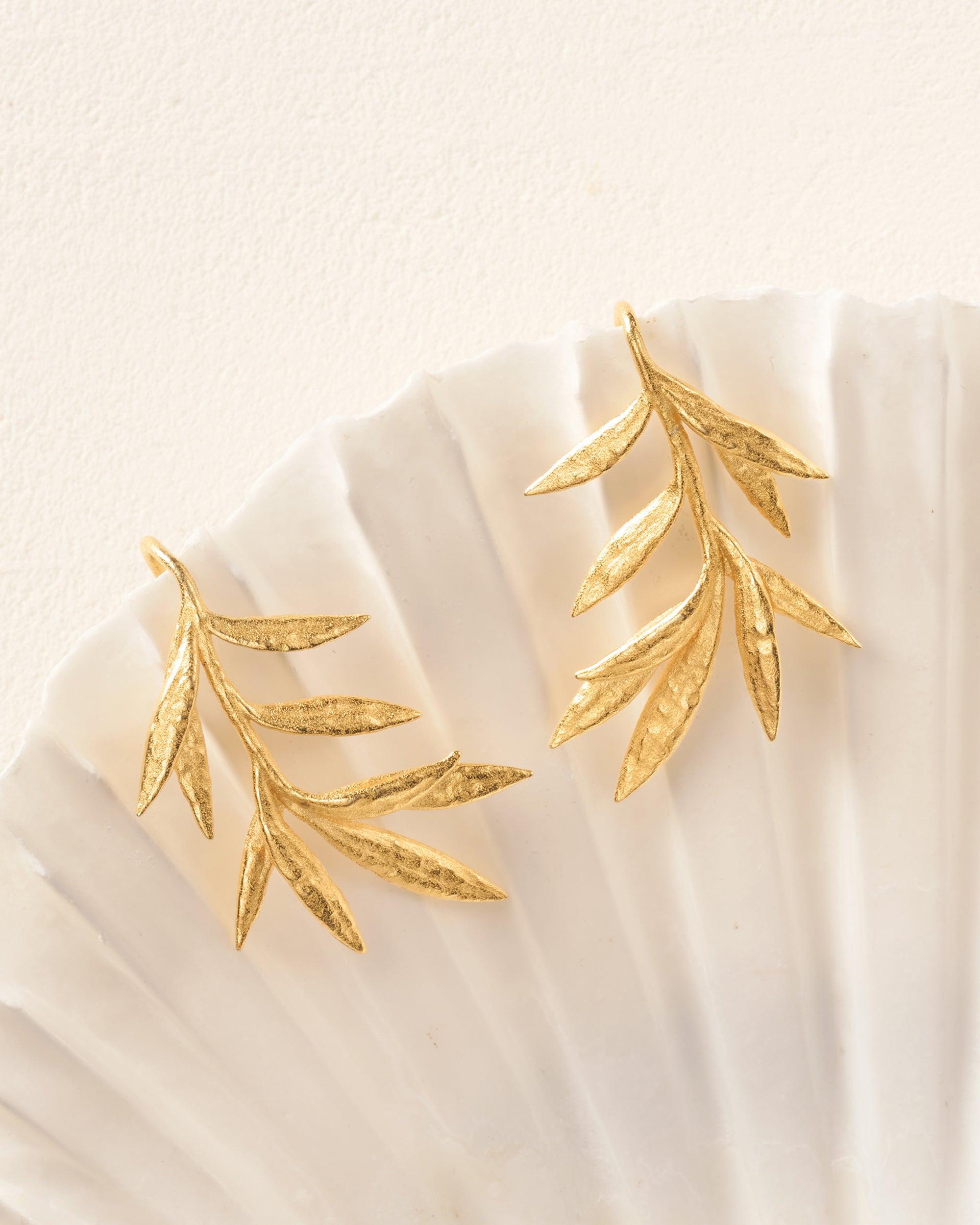 Pair of gold Elaia olive branch earrings with delicate leaves, resting on a white seashell.