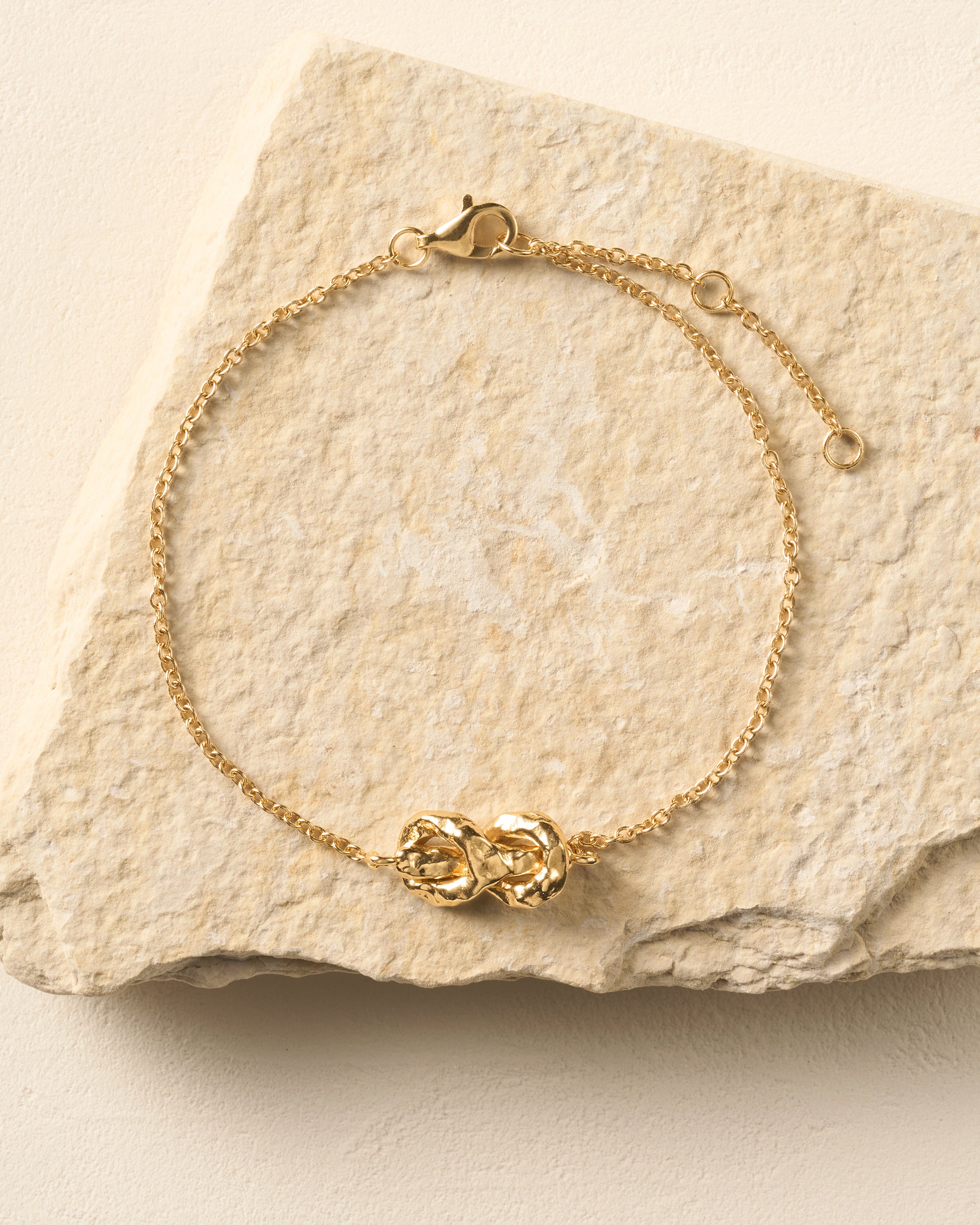 Gold Nodéa bracelet with a hammered reef knot design on a fine link chain, resting on a stone.
