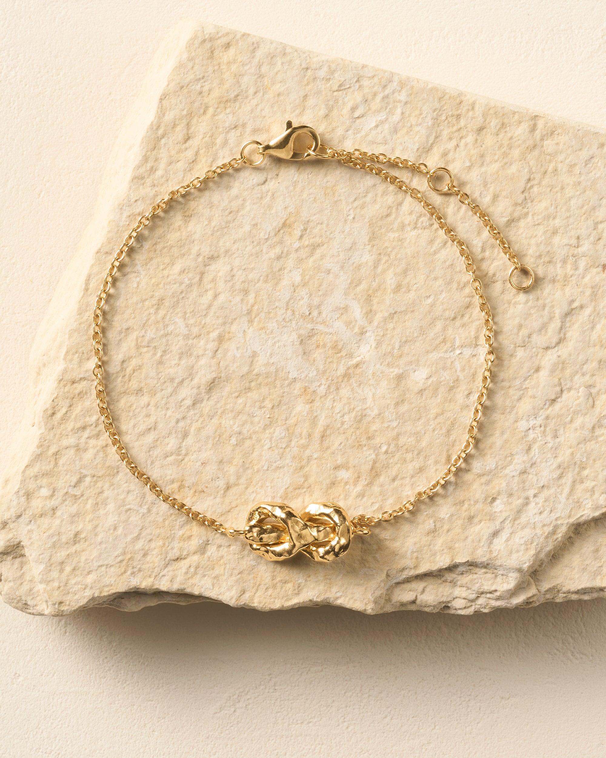 Gold Nodéa bracelet with a hammered reef knot design on a fine link chain, resting on a stone.