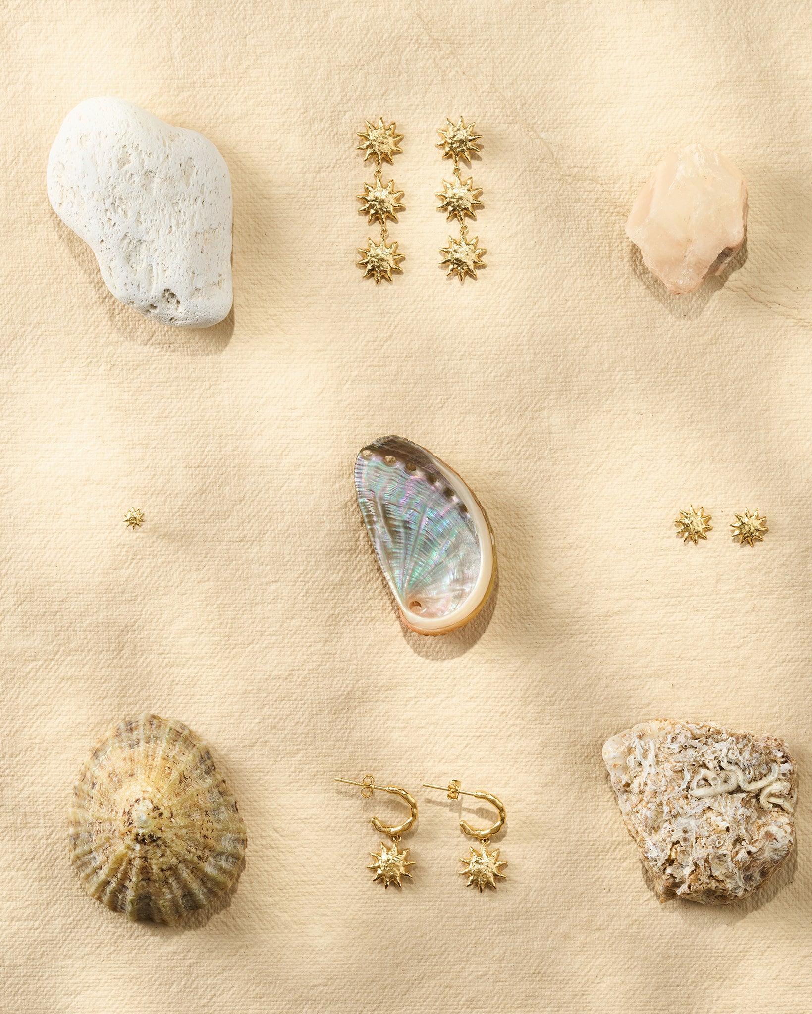 Gold Solé Stud Earrings with sculpted, hammered suns, displayed with shells and stones on a beige surface.