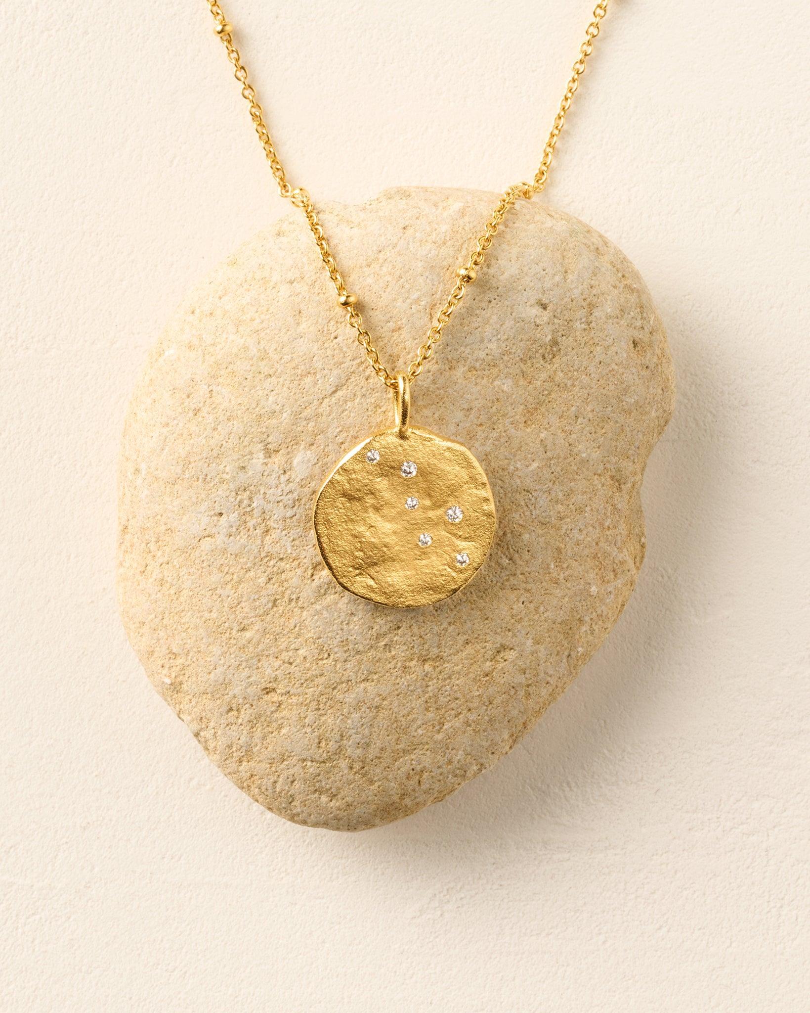 Collier Lumoria : pendentif disque rond doré à la finition texturée, incrusté de petites pierres blanches, posé sur une pierre claire.