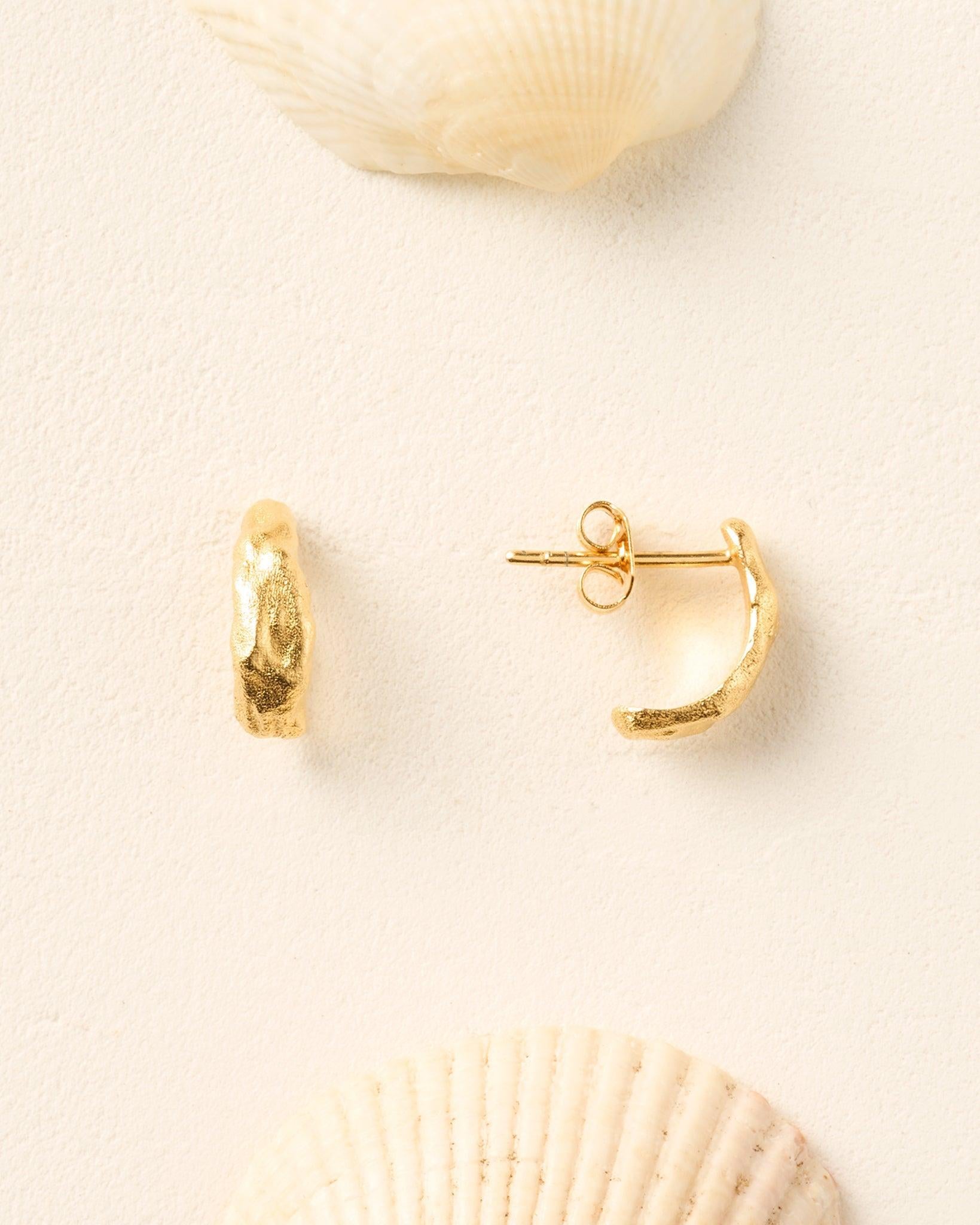 Analia small gold half-hoop earrings with an irregular, textured finish, displayed on a cream background with seashells.