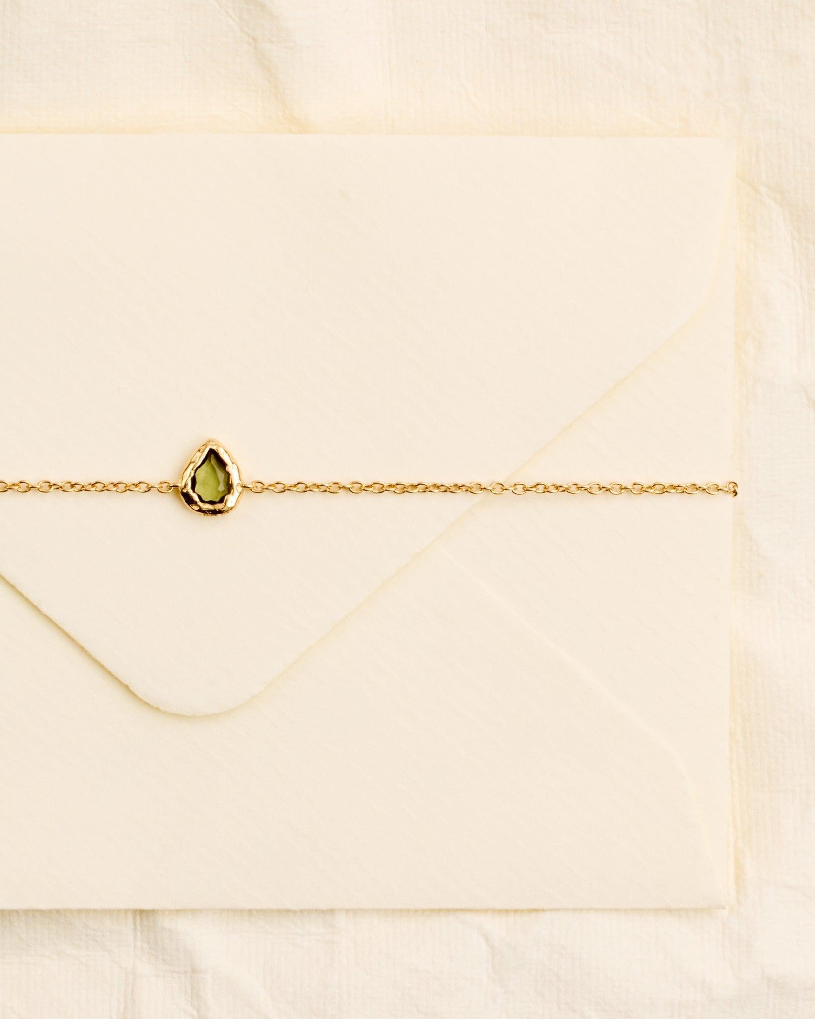 Lysia Bracelet: Gold chain with a small, textured gold drop motif holding a dark-green stone, on an envelope.
