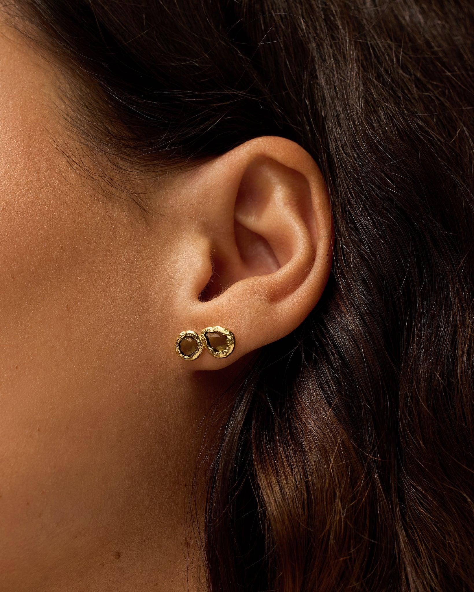 Lysiana earrings: textured gold stud with dark-green stone, worn on an ear.