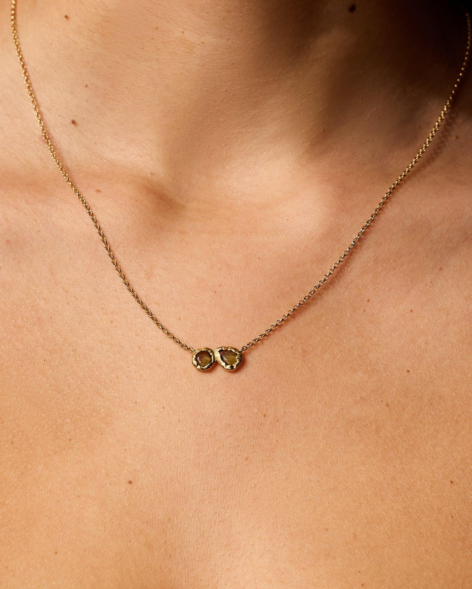 Collier Lysiana avec deux pendentifs dorés texturés, chacun orné d'une pierre vert foncé, porté au cou d'une mannequin.