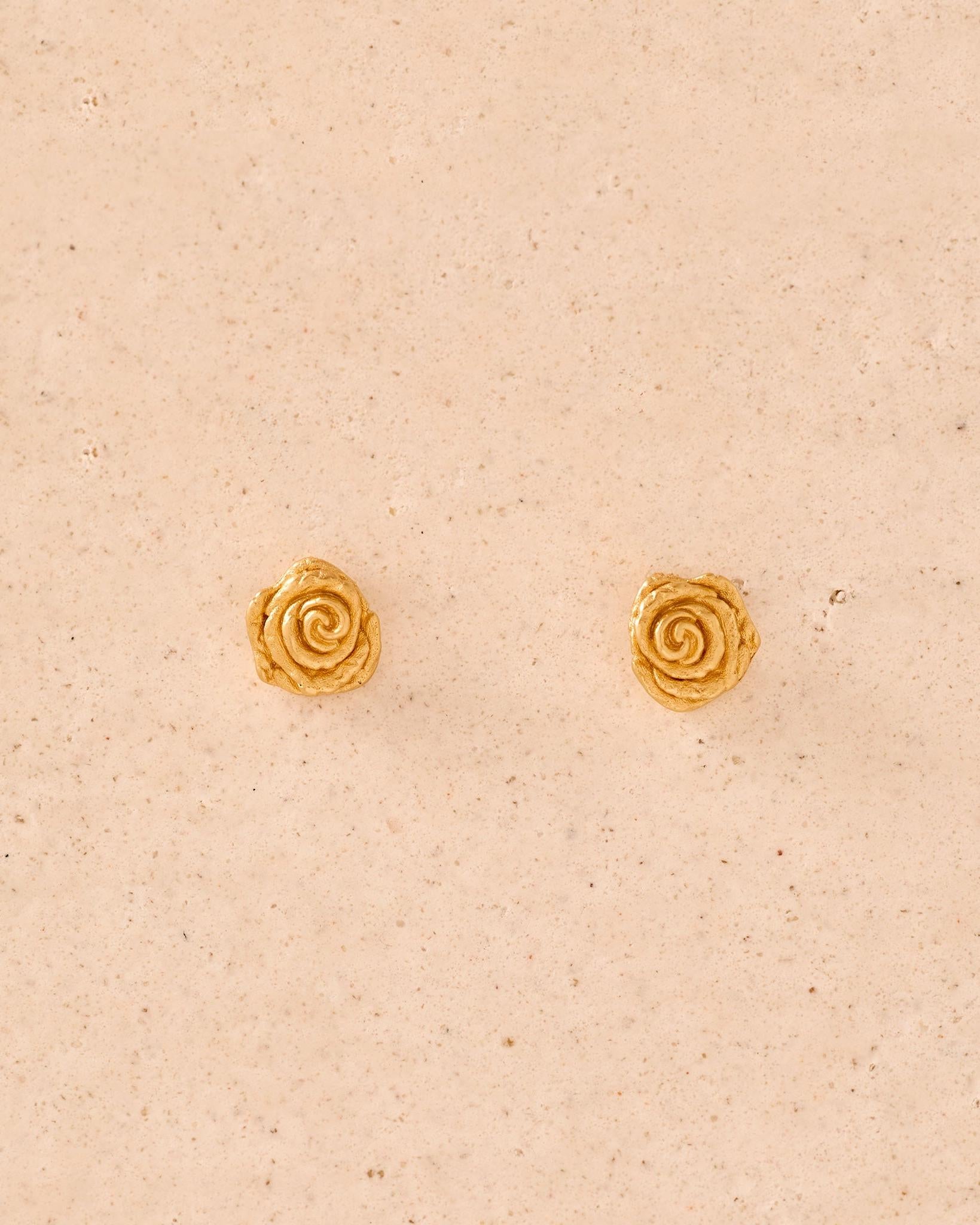 Puces d'oreille dorées en forme de roses miniatures aux pétales texturés et sculptés, sur fond beige neutre.