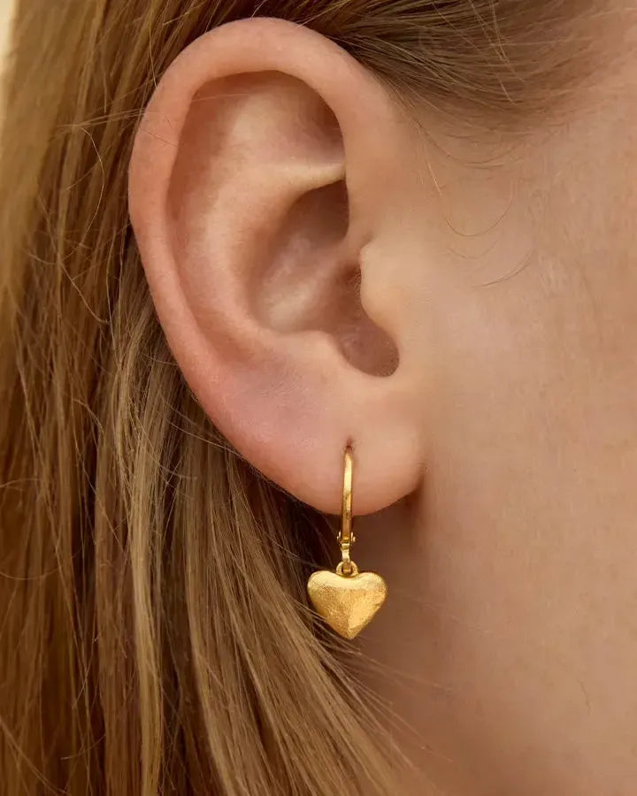 Close-up of a gold Amá earring: small, textured heart charm on a delicate hoop, worn on an ear.