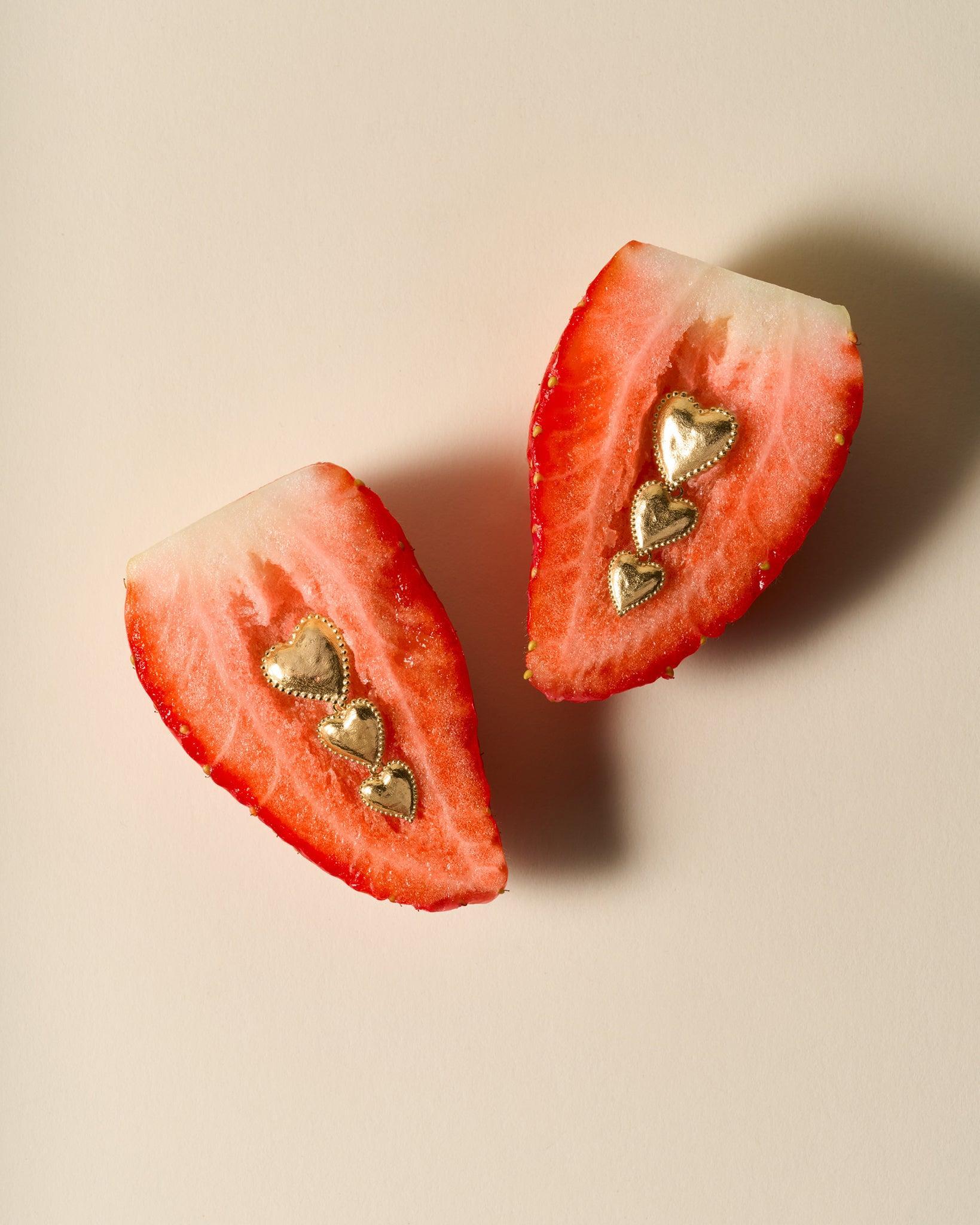 Amori Trio gold heart drop earrings with three stacked, hammered hearts and beaded halos, displayed on strawberry halves.