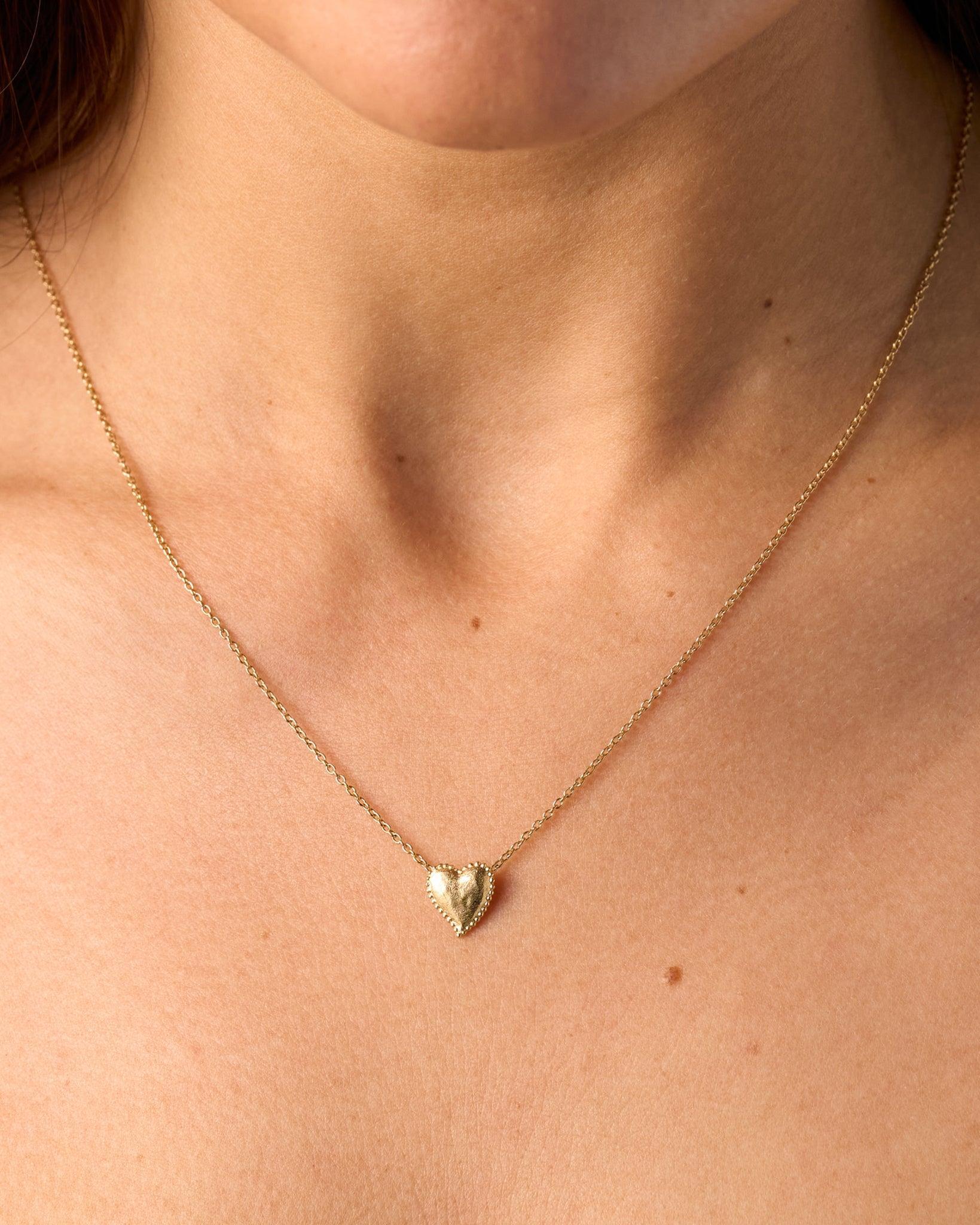 Collier Amori doré avec un pendentif cœur martelé et une auréole de perles sur une chaîne fine, porté sur la poitrine d'une personne.