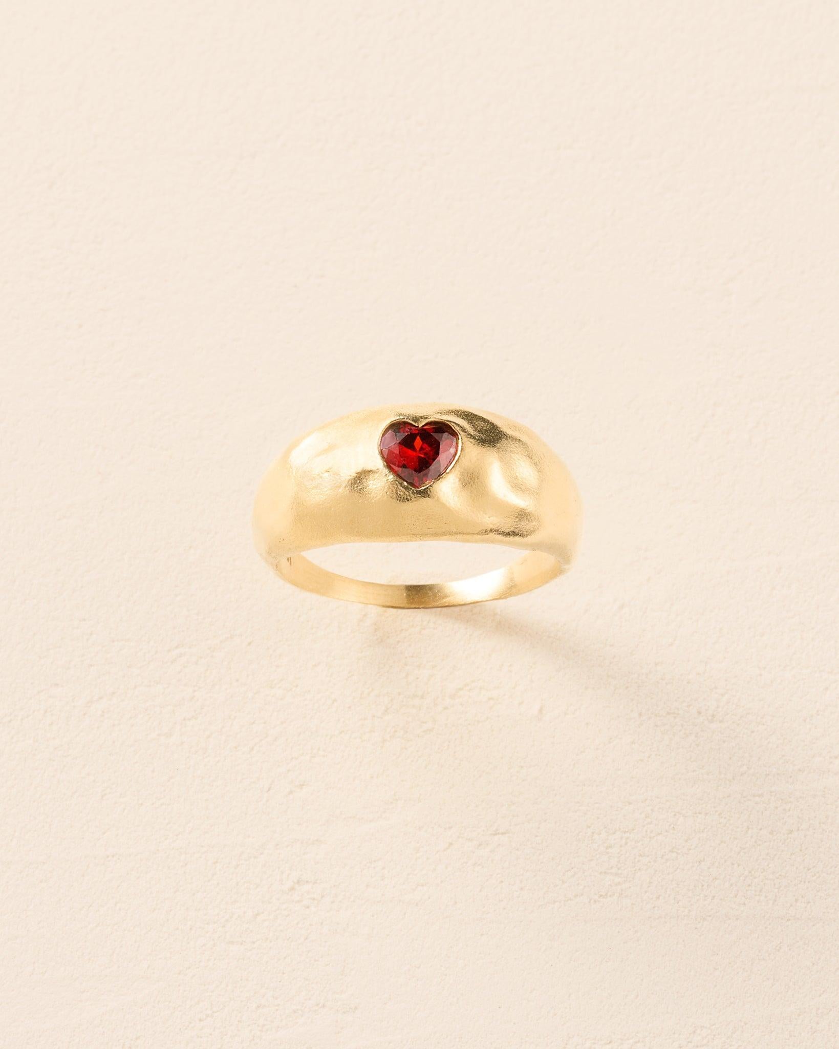 Luzia Red Ring: Gold domed ring with a deep red heart-shaped stone on a hammered surface.