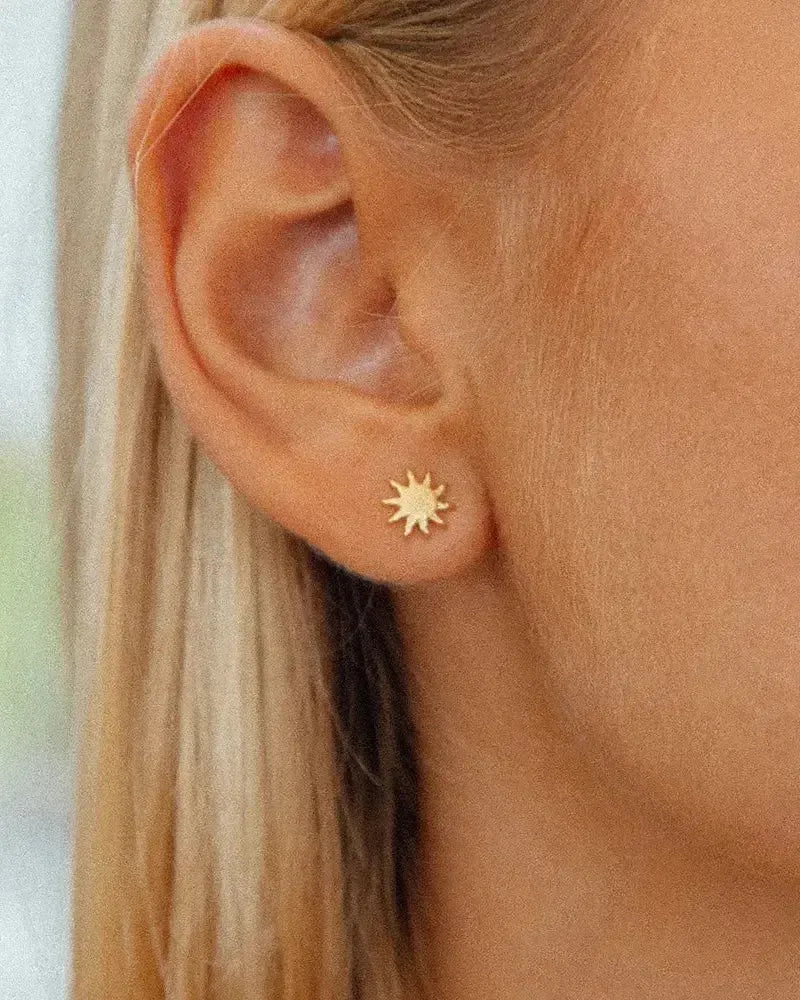 Close-up of a woman's ear wearing a small, golden sun-shaped stud earring.