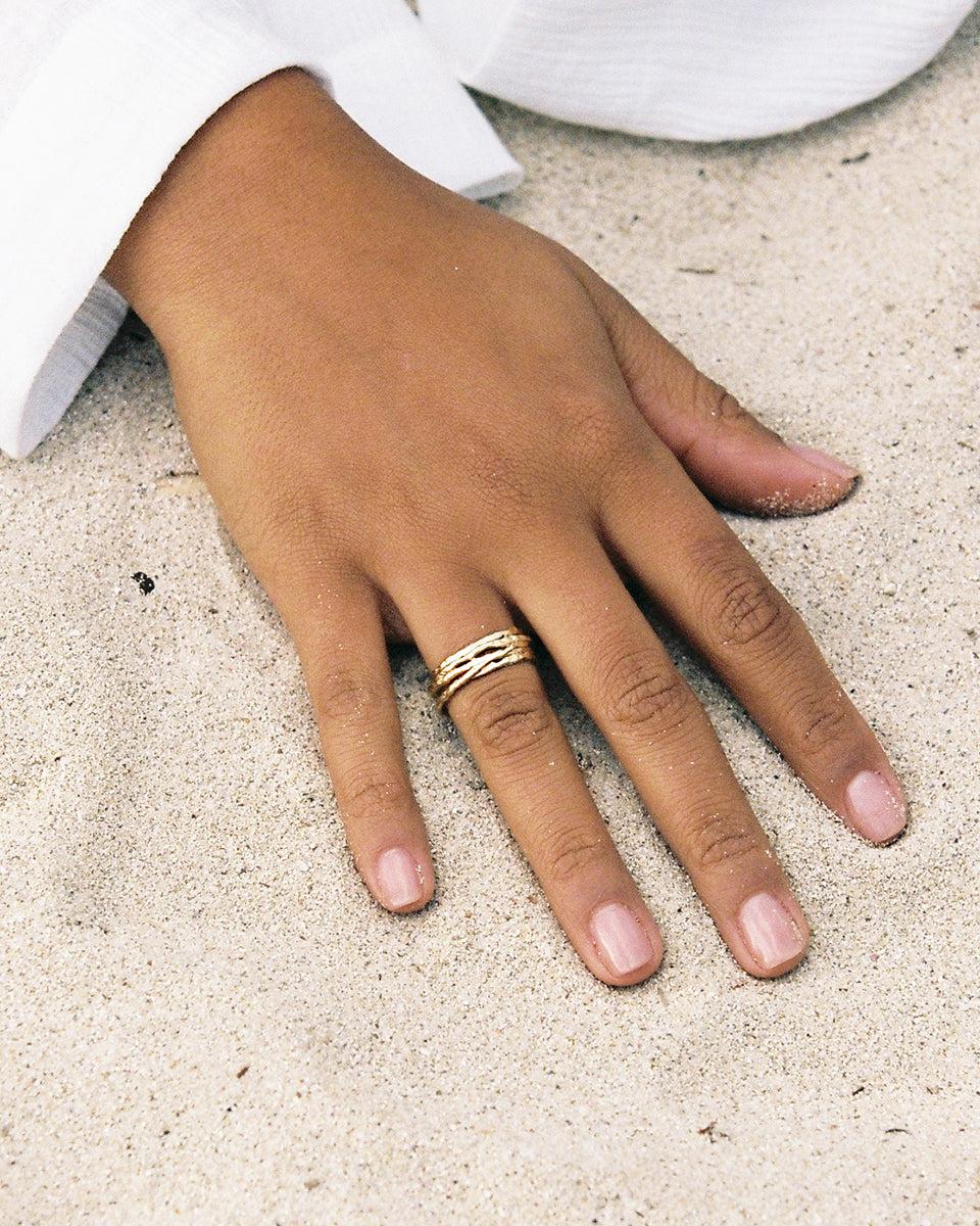 Hand on sand wearing a gold Coral ring with organic, textured lines.