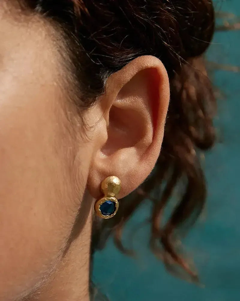 Gold Petra stud earring with a deep night blue stone on a woman's ear.