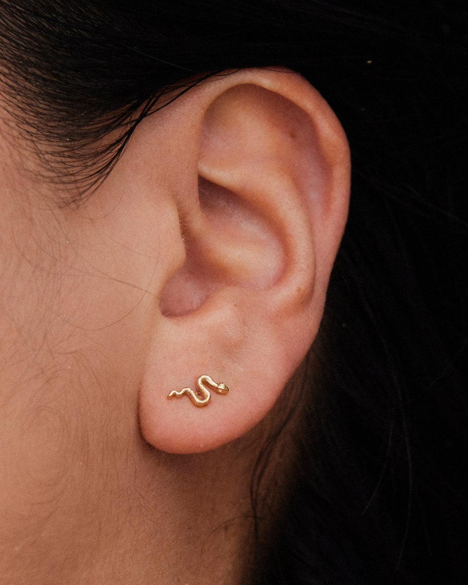 Minimalistischer, goldfarbener Schlangen-Ohrstecker, am Ohrläppchen getragen, mit zartem, geschwungenem Schlangendesign.