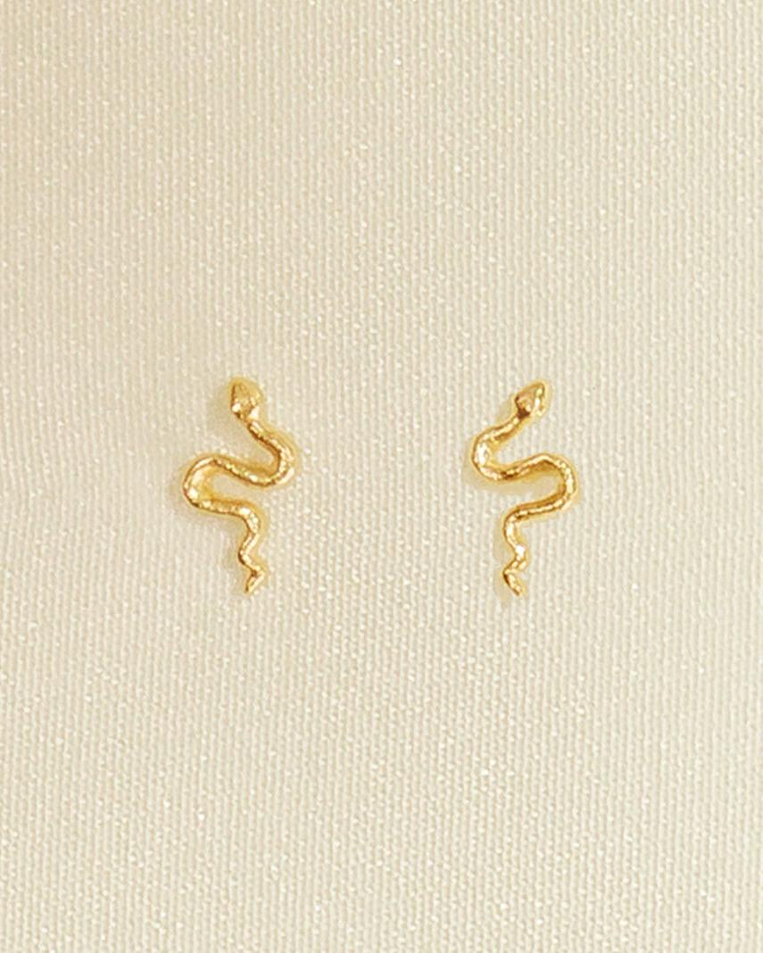 Petites puces d'oreille en forme de serpent dorées à la finition texturée, sur fond en tissu neutre.