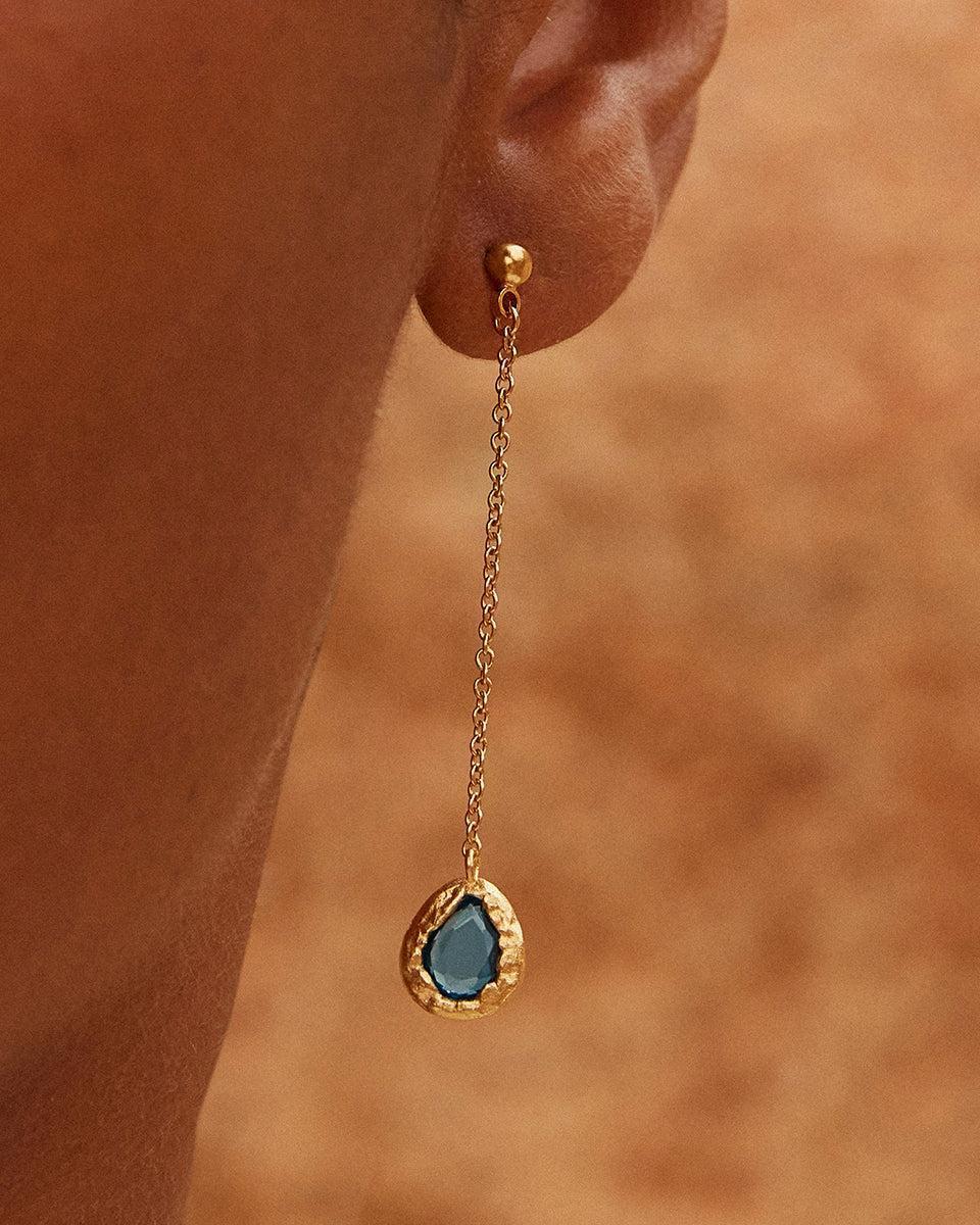 Lysia long gold earrings with thin chains and drop-shaped blue stone pendants in textured gold.