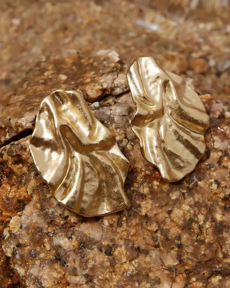 Pair of gold Batla earrings with a soft petal, fabric-like texture, resting on brown rocks.