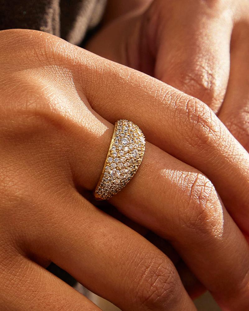 Breiter, gewölbter, goldfarbener Ring mit einer im Pavé gefassten Oberfläche aus funkelnden, weißen Steinen, an einem Finger getragen.