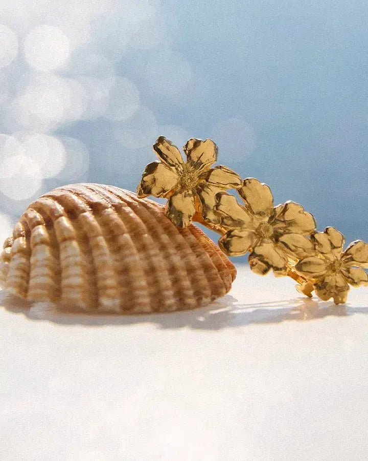 Pinza para el pelo dorada Rosaria con tres flores delicadas en flor, apoyada sobre una concha.