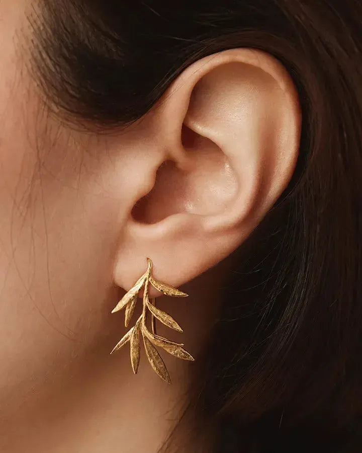 Gold Elaia olive branch earrings, delicate and curved, worn on an ear.