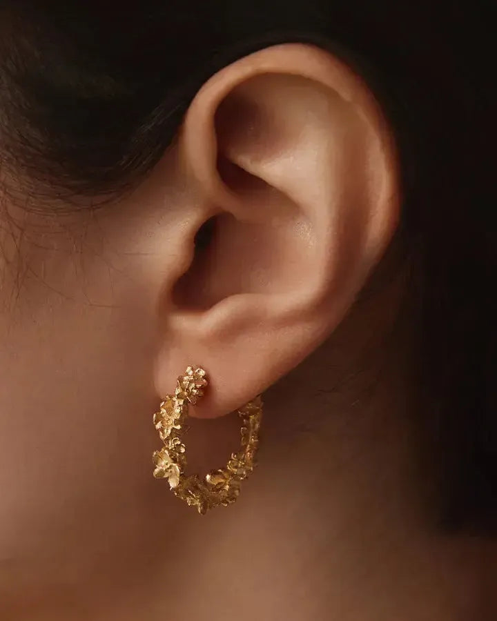 Gold Kèpos hoop earring with intricate floral garland design on an ear.