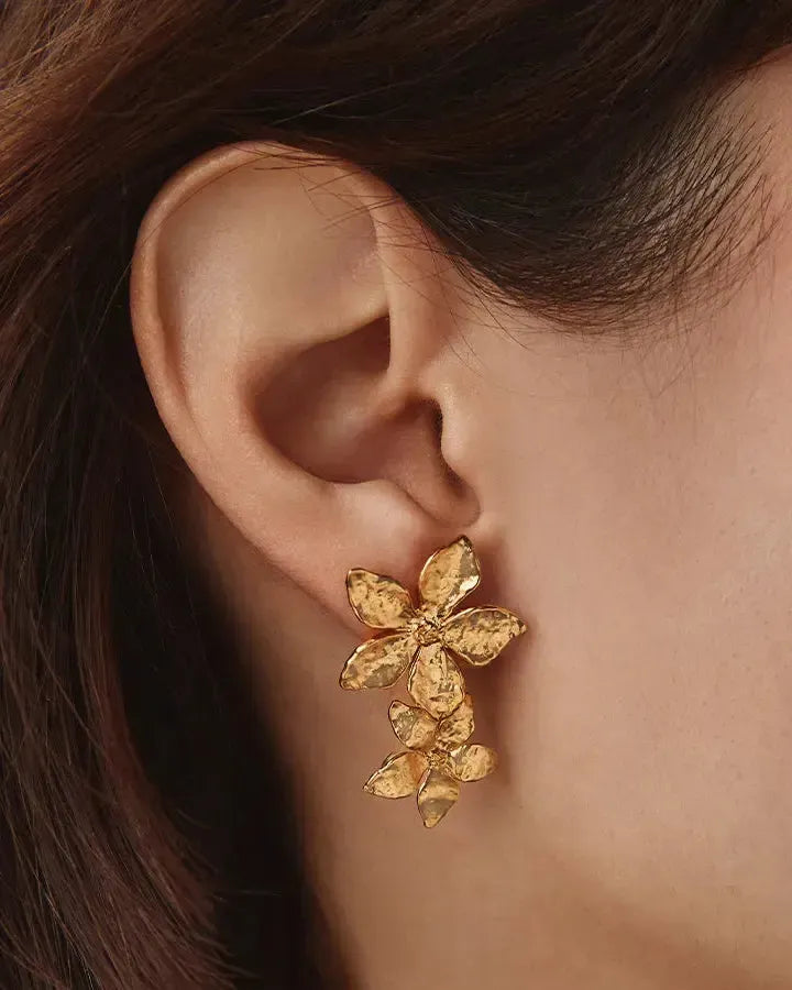 Gold hammered floral drop earrings with two five-petal flowers on a woman's ear.