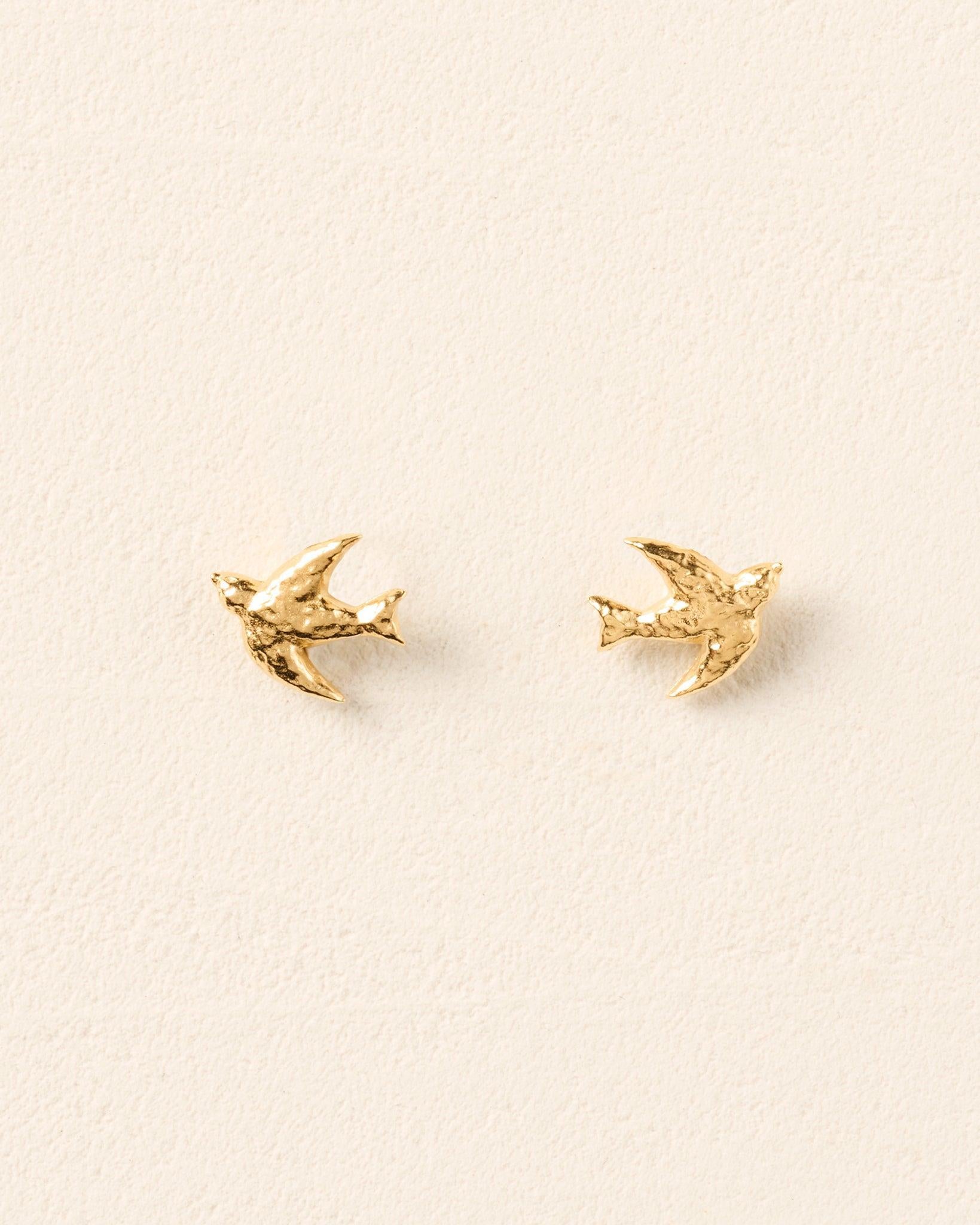 Gold Avila stud earrings shaped like flying birds with sculpted, textured wings on a light cream background.