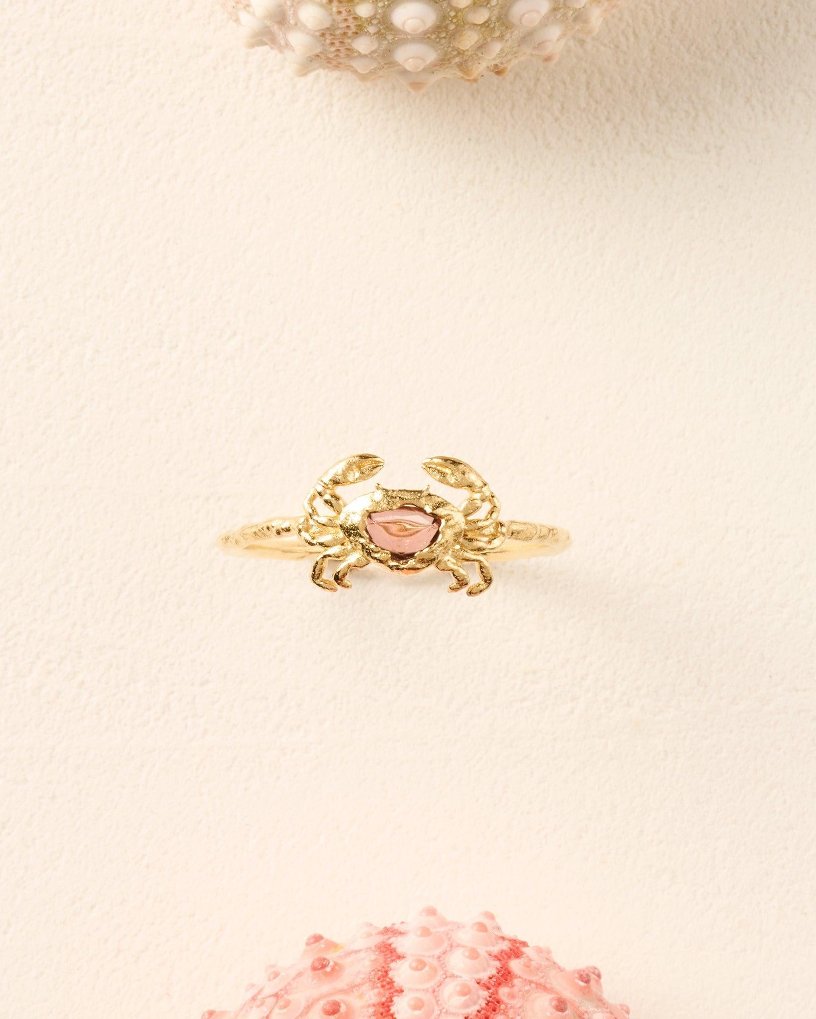 Textured gold crab ring featuring a central red stone on a thin band, set against a light cream background.