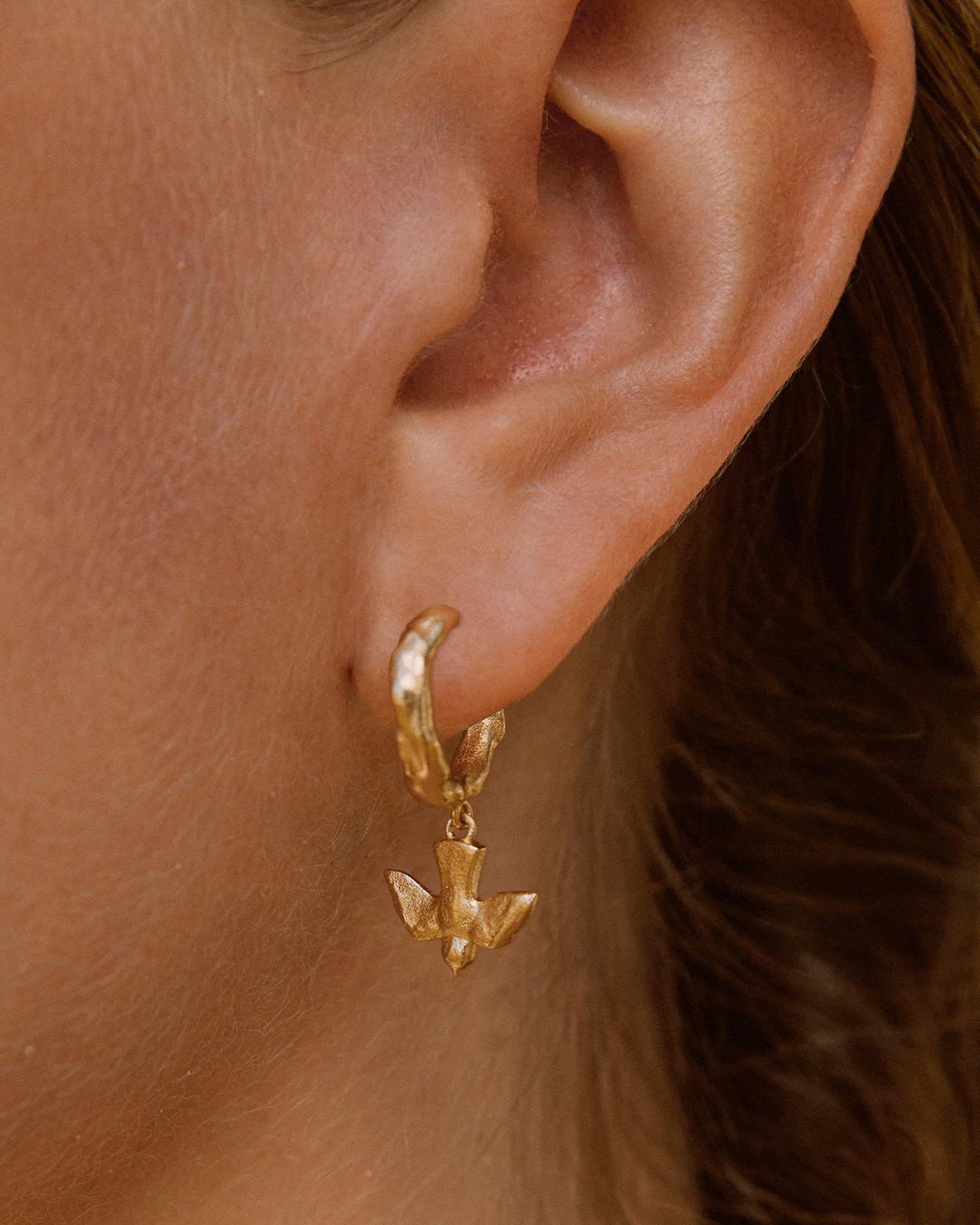 Gold Mara hoop earrings featuring a small, detailed flying bird pendant with outstretched wings on a textured band.