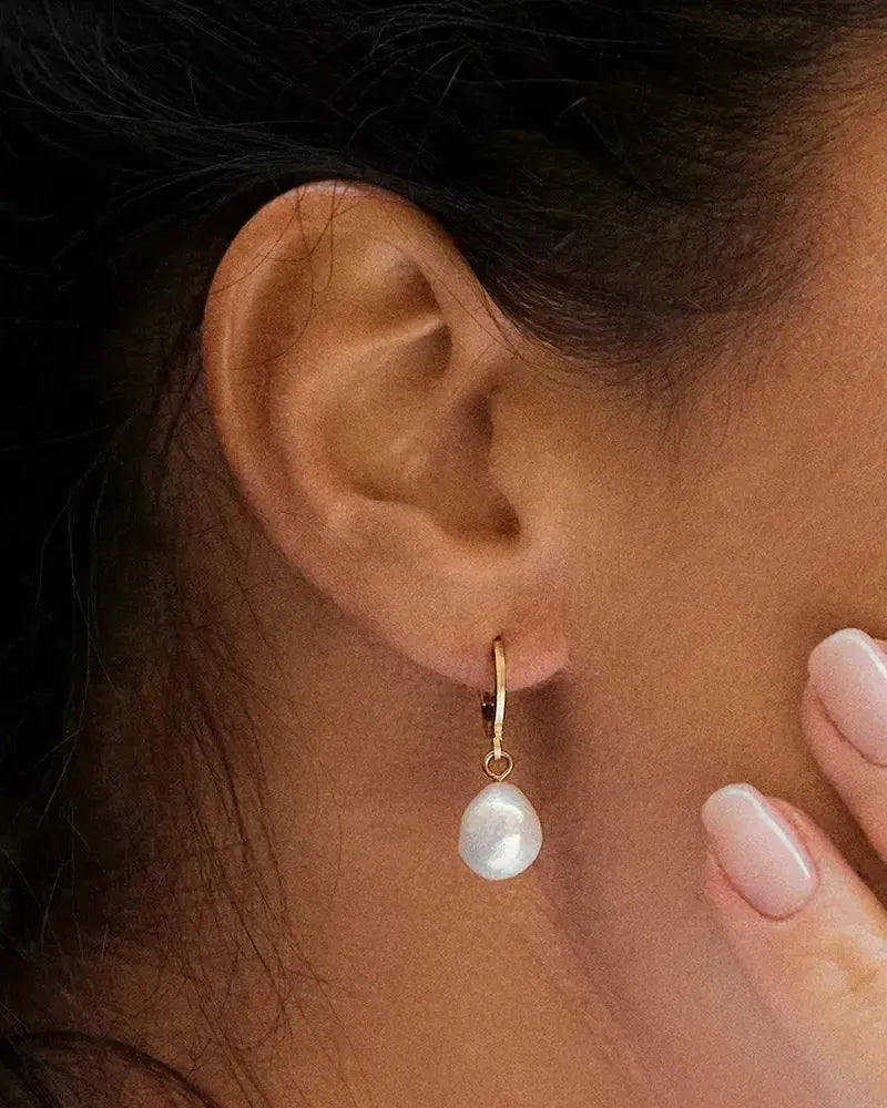 Gold hoop earring with a dangling white baroque pearl and small red gem on a woman's ear.