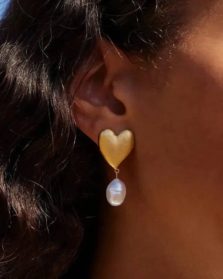 Pendientes Ángel dorados en forma de corazón con una perla barroca blanca colgante, llevados en una oreja.