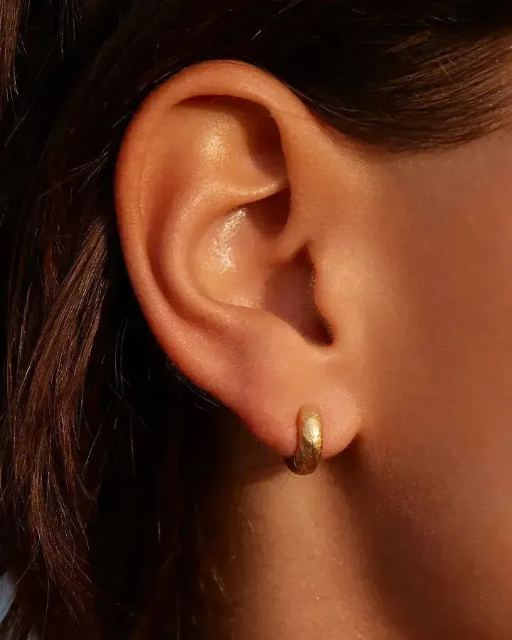 Close-up of a model's ear wearing a single gold Anilla Mini hammered hoop earring.