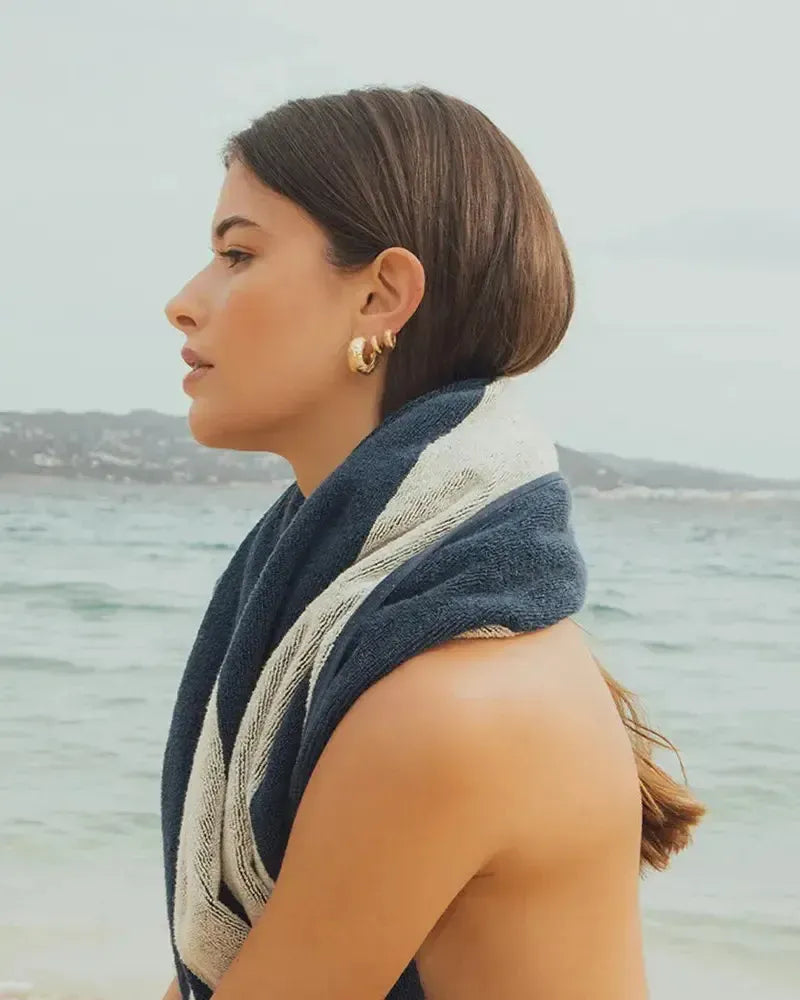 Woman wearing Anilla Mini gold hammered hoop earrings with a blue and white striped towel by the ocean.