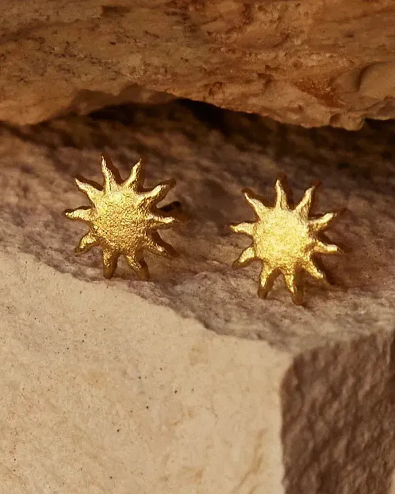 Par de pendientes de botón de sol dorados texturizados con rayos radiantes, posados sobre una piedra de color claro.