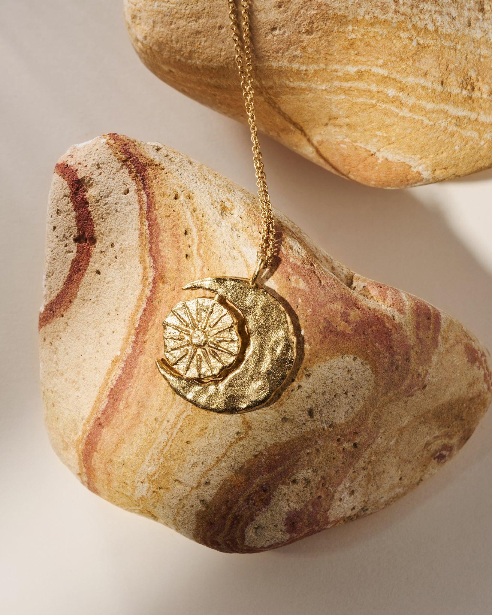 Collier Phébé doré à croissant de lune avec pendentif soleil rotatif, présenté sur des roches texturées.