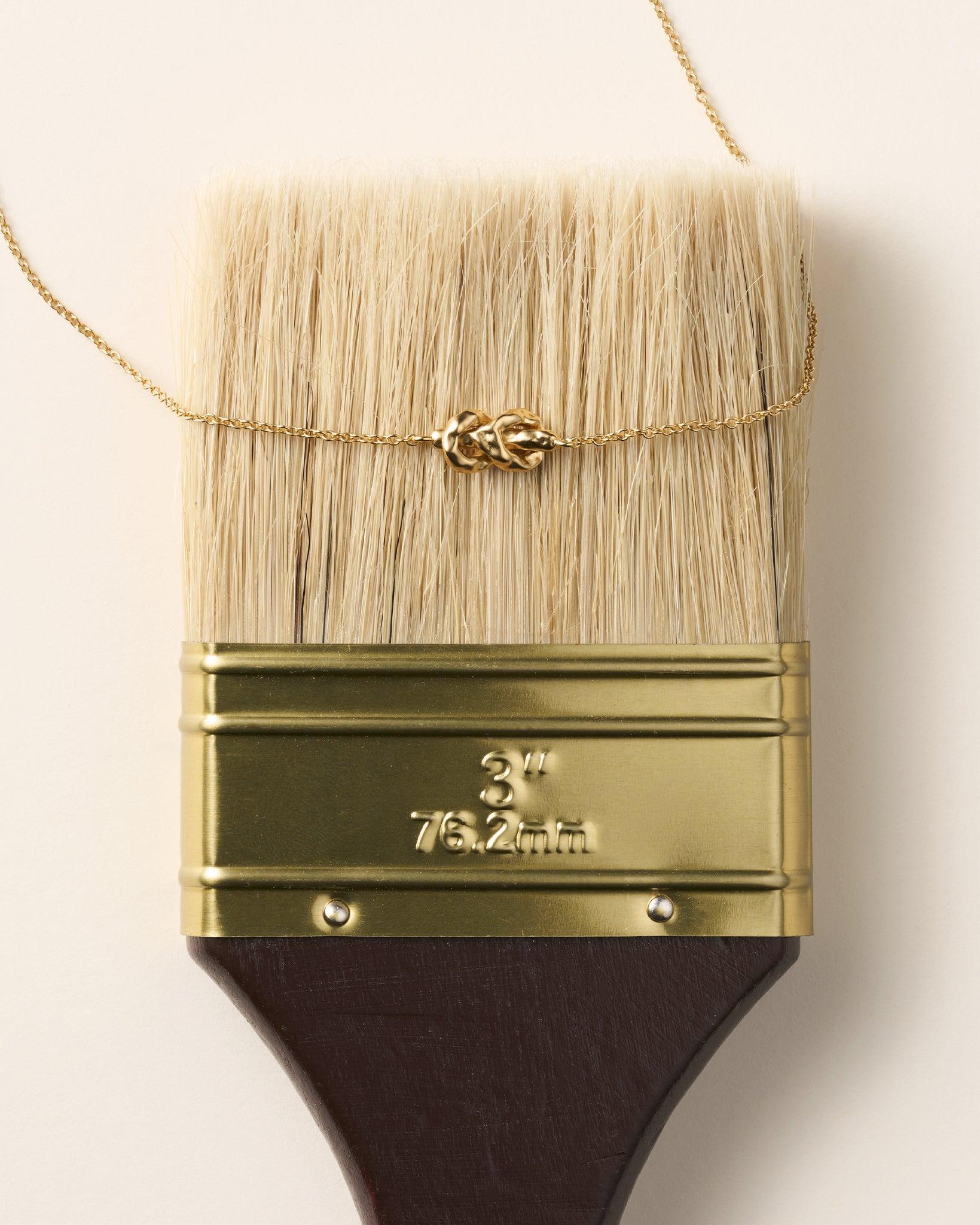 Gold Nodéa necklace with hammered reef knot pendant on a fine chain, resting on a paintbrush.