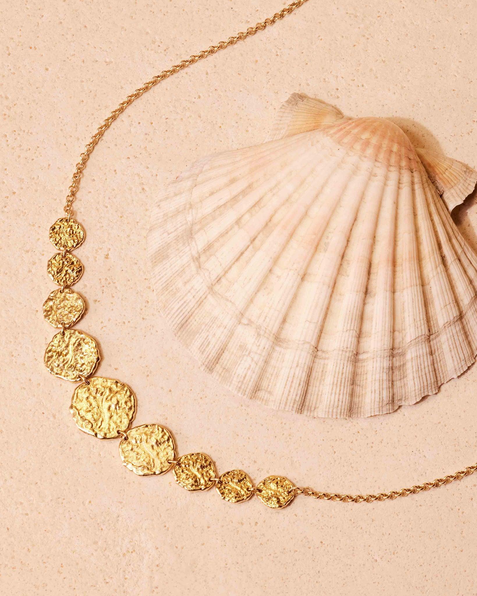 Collier Cassia dégradé avec 9 disques dorés martelés, façon bijou à pièces anciennes, à côté d'un coquillage.
