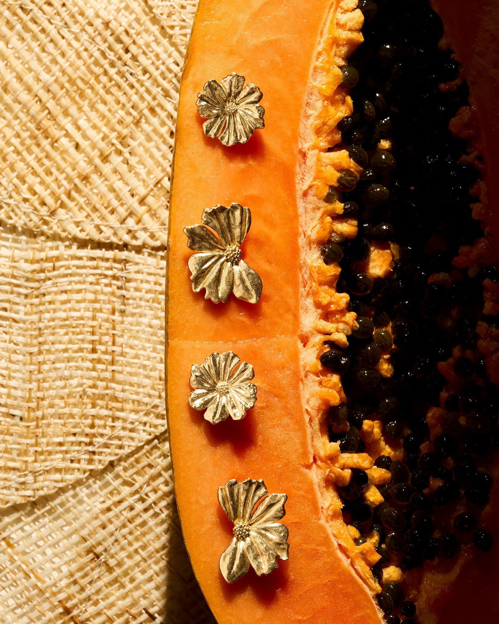 Gold Mona floral earrings with sculpted, layered petals resting on a halved papaya against a textured woven background.