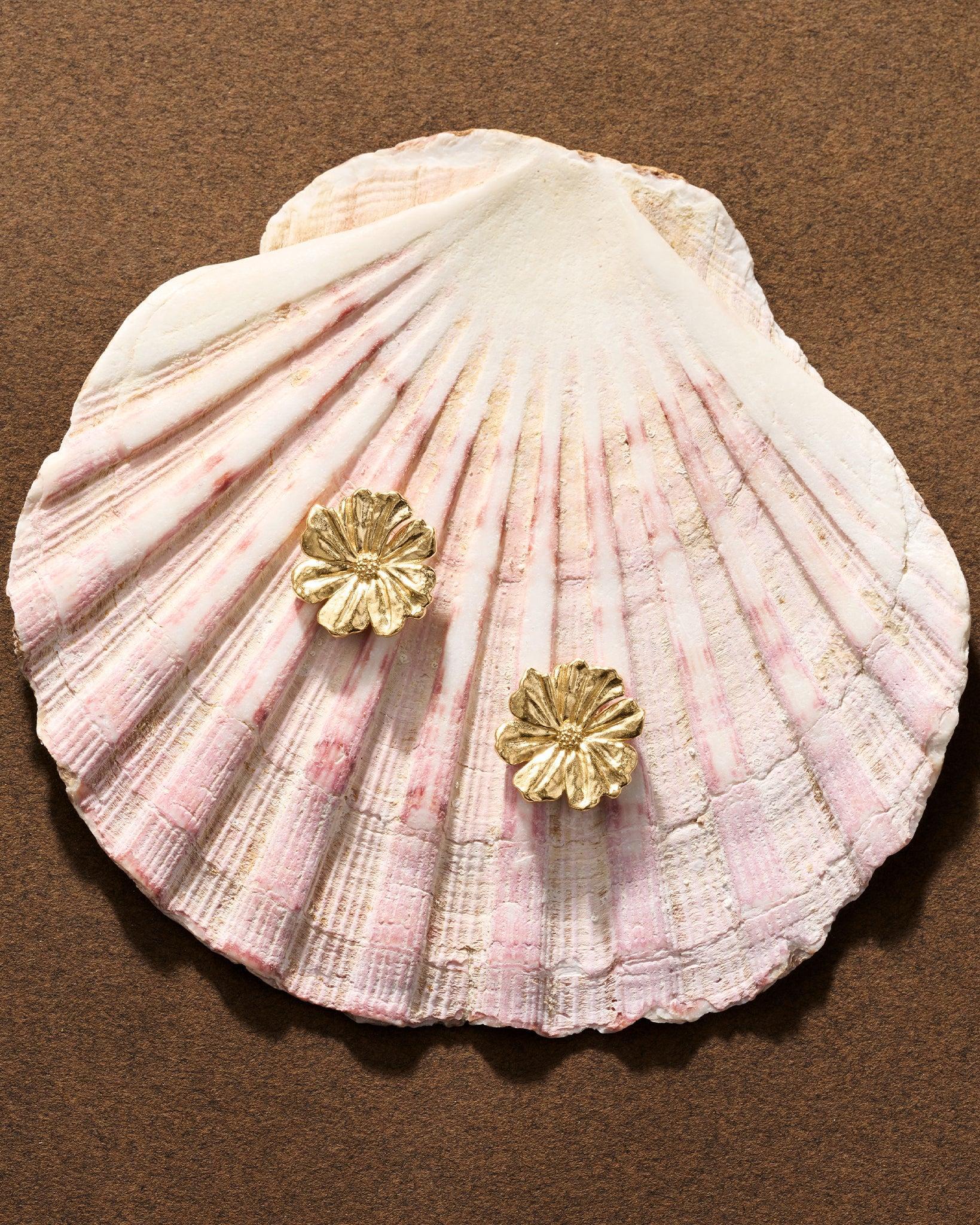 Gold Mona floral earrings with sculpted, layered petals resting on a textured pink and white seashell.