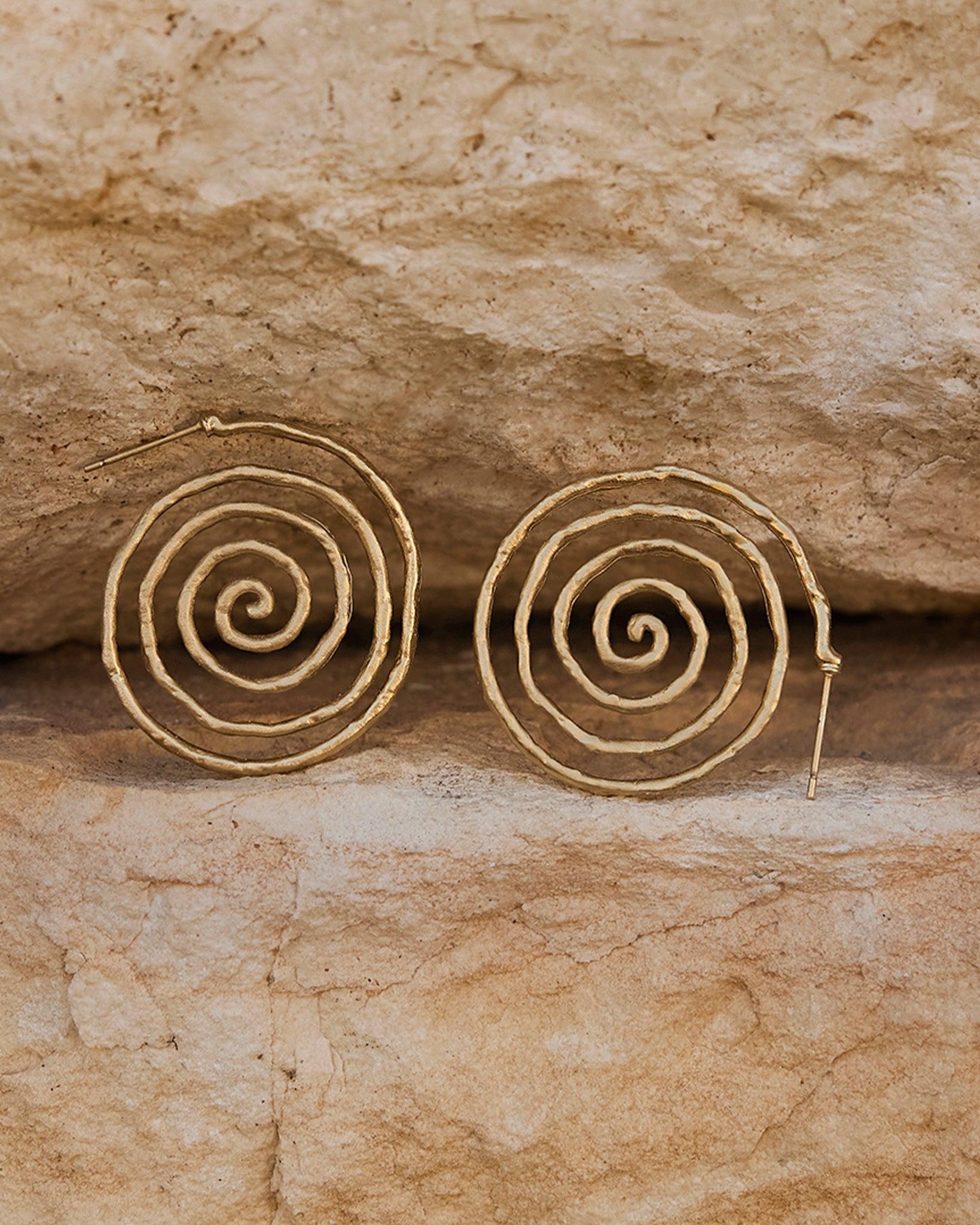 Boucles d'oreilles spirale Nacci dorées, fluides et sinueuses, sur un fond de pierre marron clair texturé.