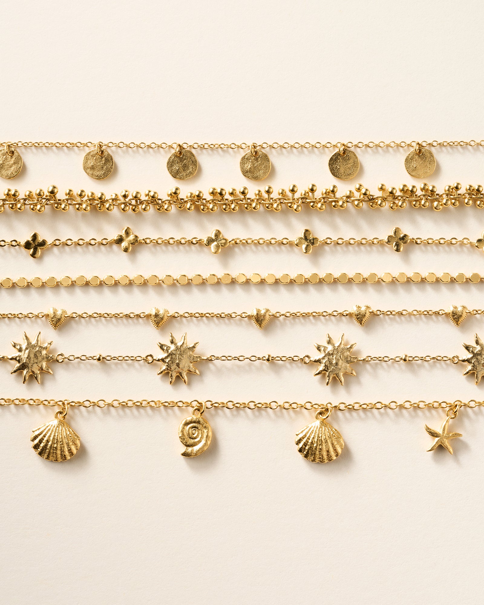 Seven delicate gold chain bracelets with various charms: flat discs, tiny beads, clover shapes, small circles, hearts, sunbursts, shells, and a starfish.
