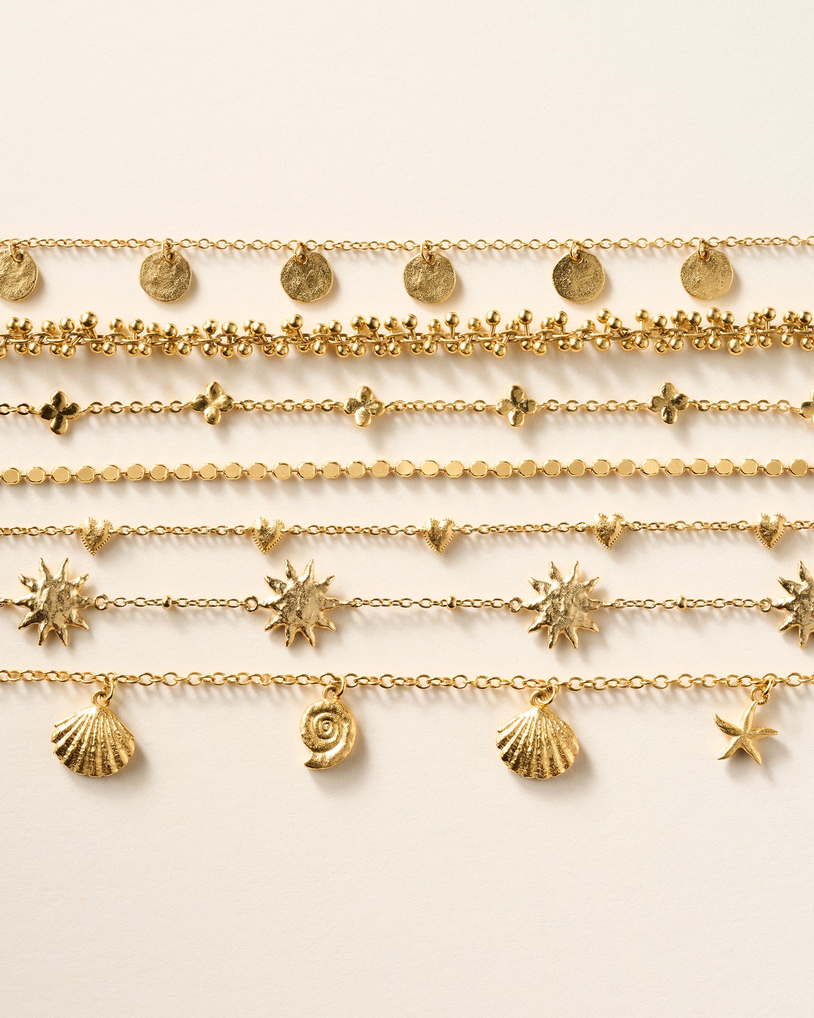 Seven delicate gold chain bracelets with various charms: flat discs, tiny beads, clover shapes, small circles, hearts, sunbursts, shells, and a starfish.