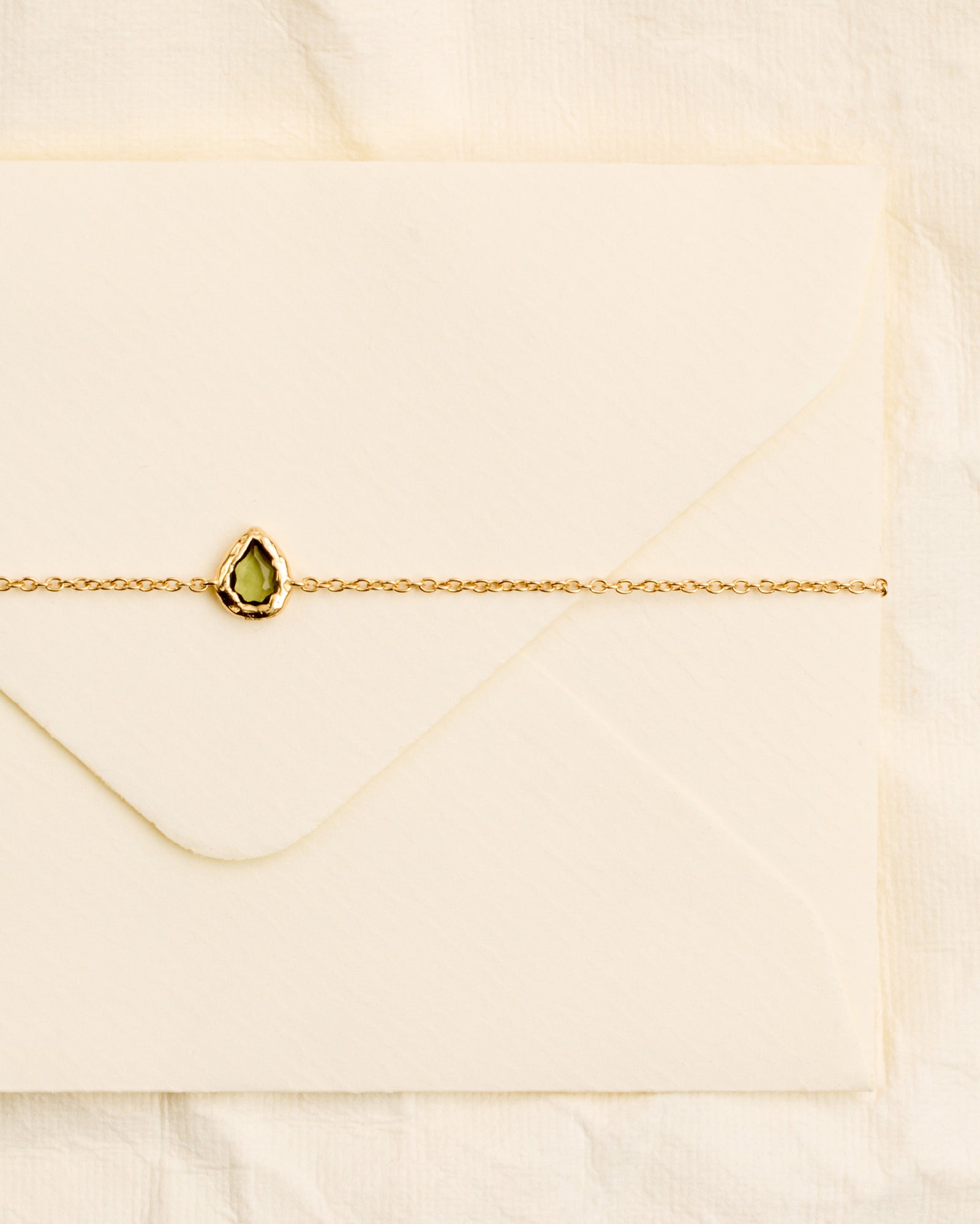 Lysia Bracelet: Gold chain with a small, textured gold drop motif holding a dark-green stone, on an envelope.
