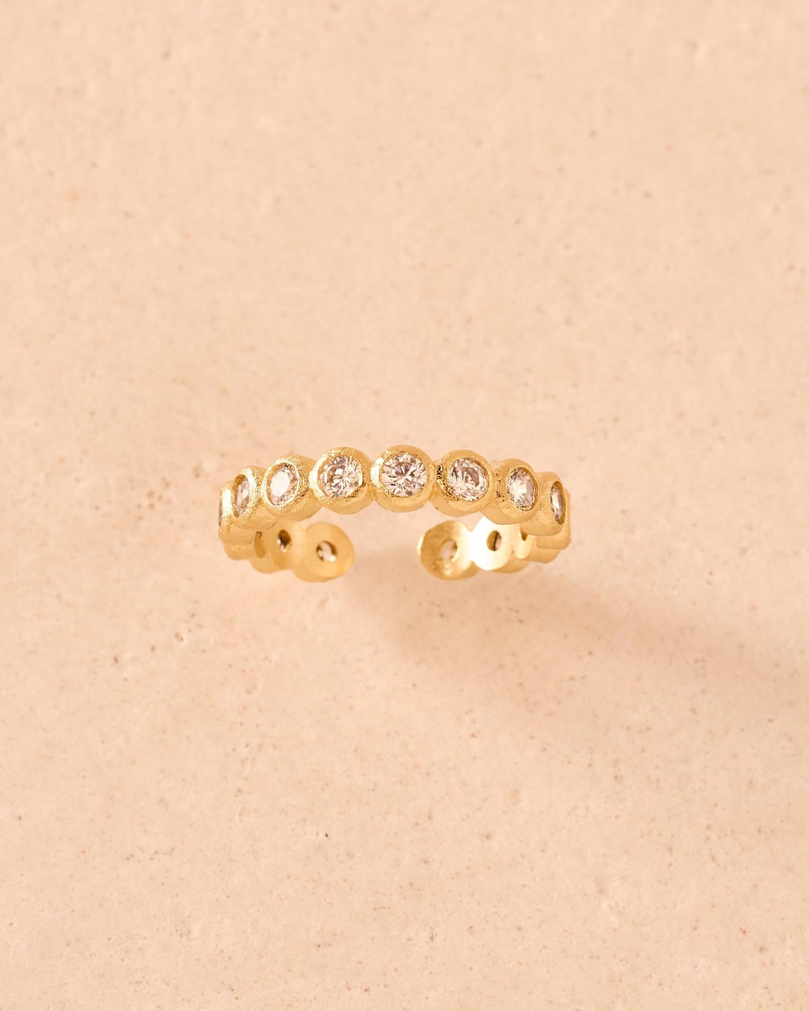 Anello dorato a fascia aperta con una fila di pietre rotonde trasparenti in un castone geometrico testurizzato a perline.