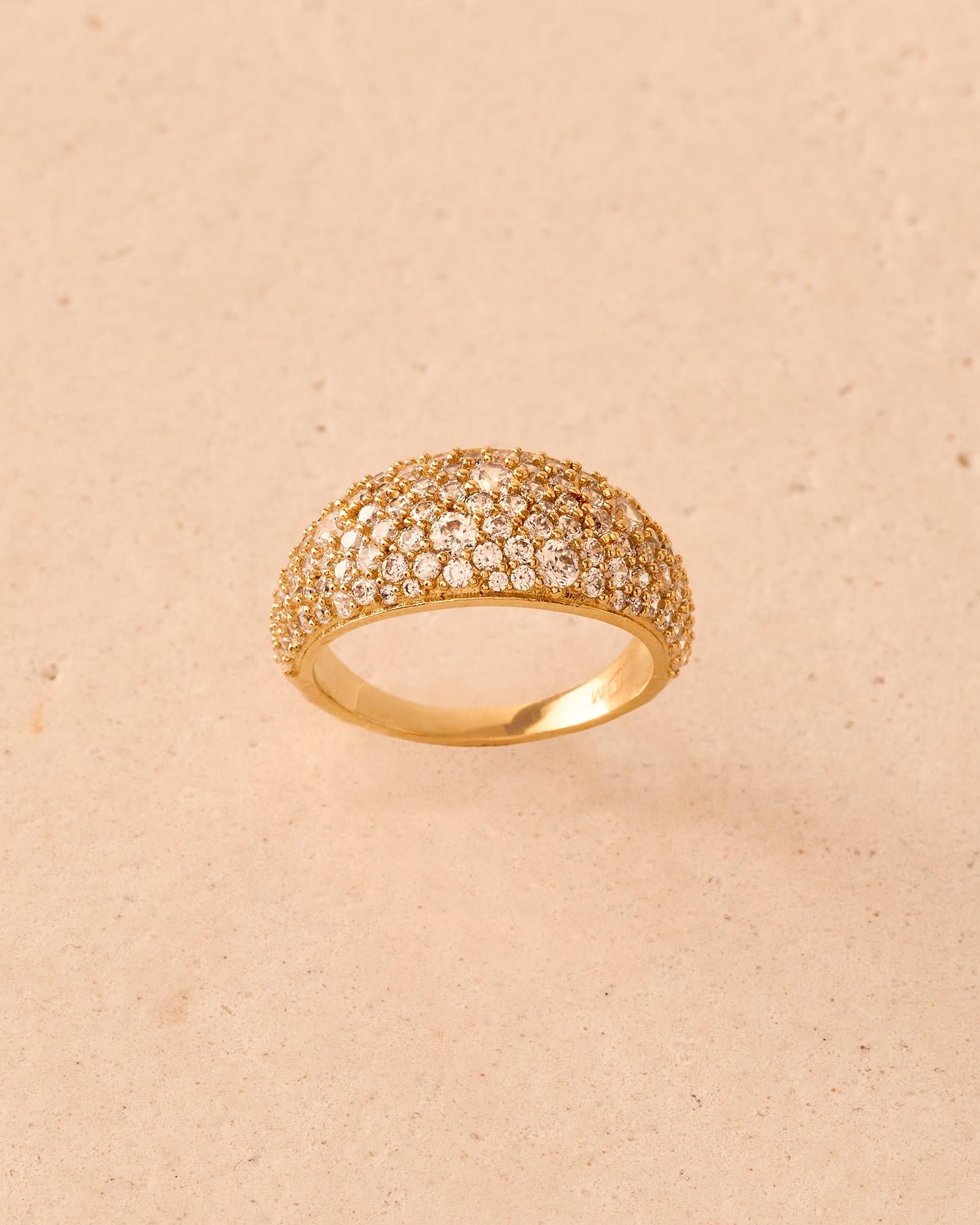 Breiter, gewölbter, goldfarbener Ring mit einer im Pavé gefassten Oberfläche aus funkelnden, weißen Steinen, vor strukturiertem, beigem Hintergrund.