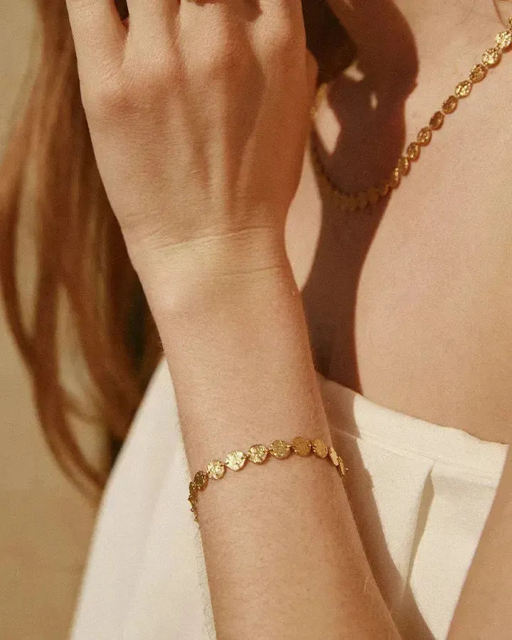 Caius Armband mit kleinen, gehämmerten goldfarbenen Scheiben am Handgelenk einer Frau, erinnert an antiken Münzschmuck.