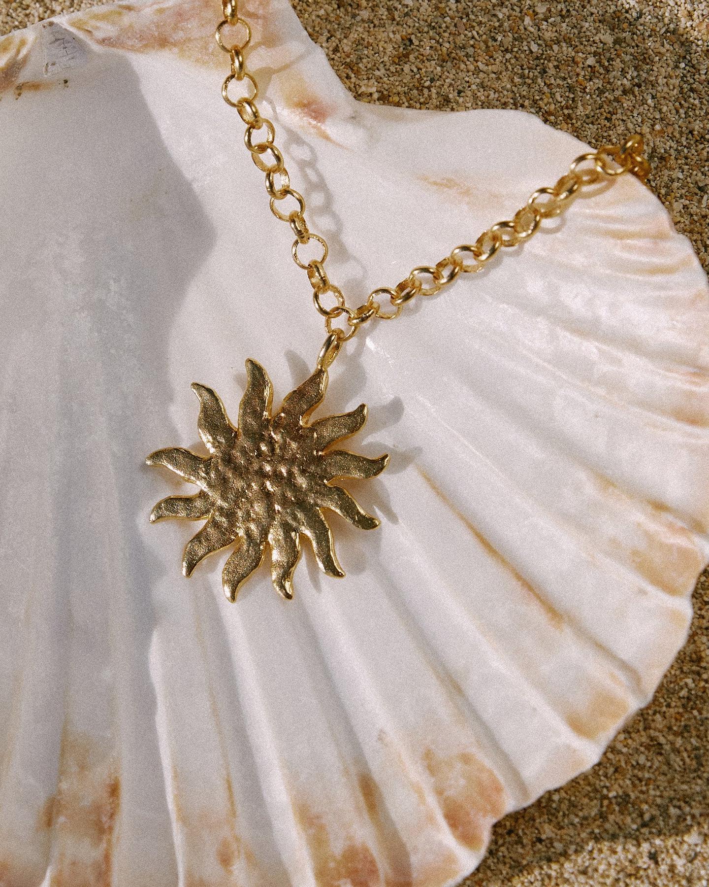Helio gold hammered sun pendant necklace with wavy rays and rounded link chain on a seashell.