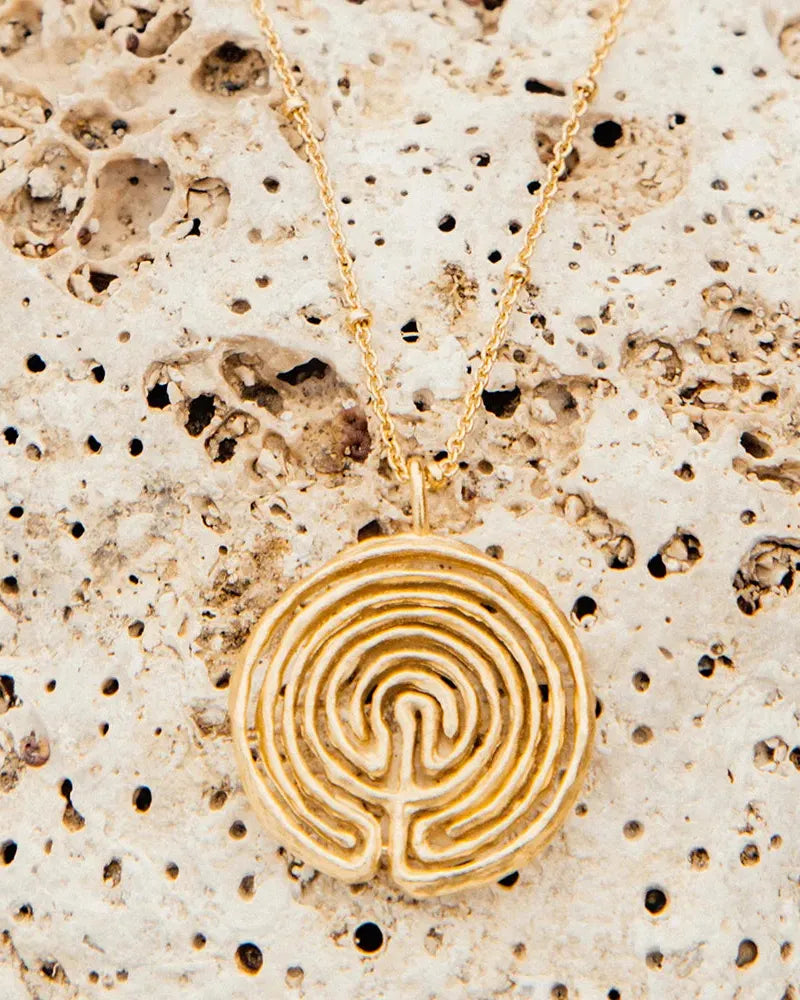Gold Knossos necklace with a circular labyrinth pendant and delicate beaded chain on a textured stone.